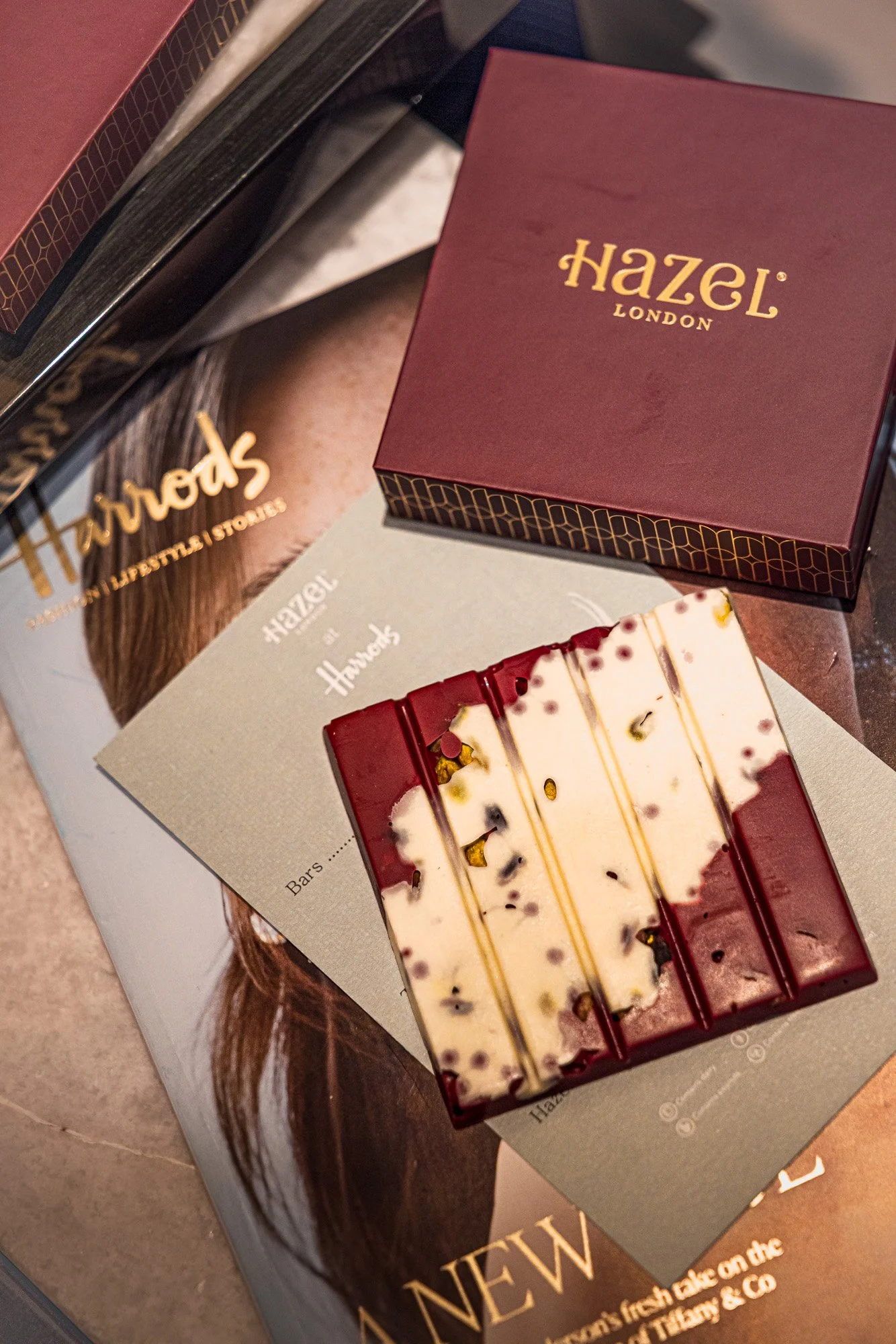 Commercial product photograph of Hazel London chocolate bars and burgundy packaging arranged with printed promotional material, showing the product range, branding and premium presentation for retail in Harrods or campaign use.