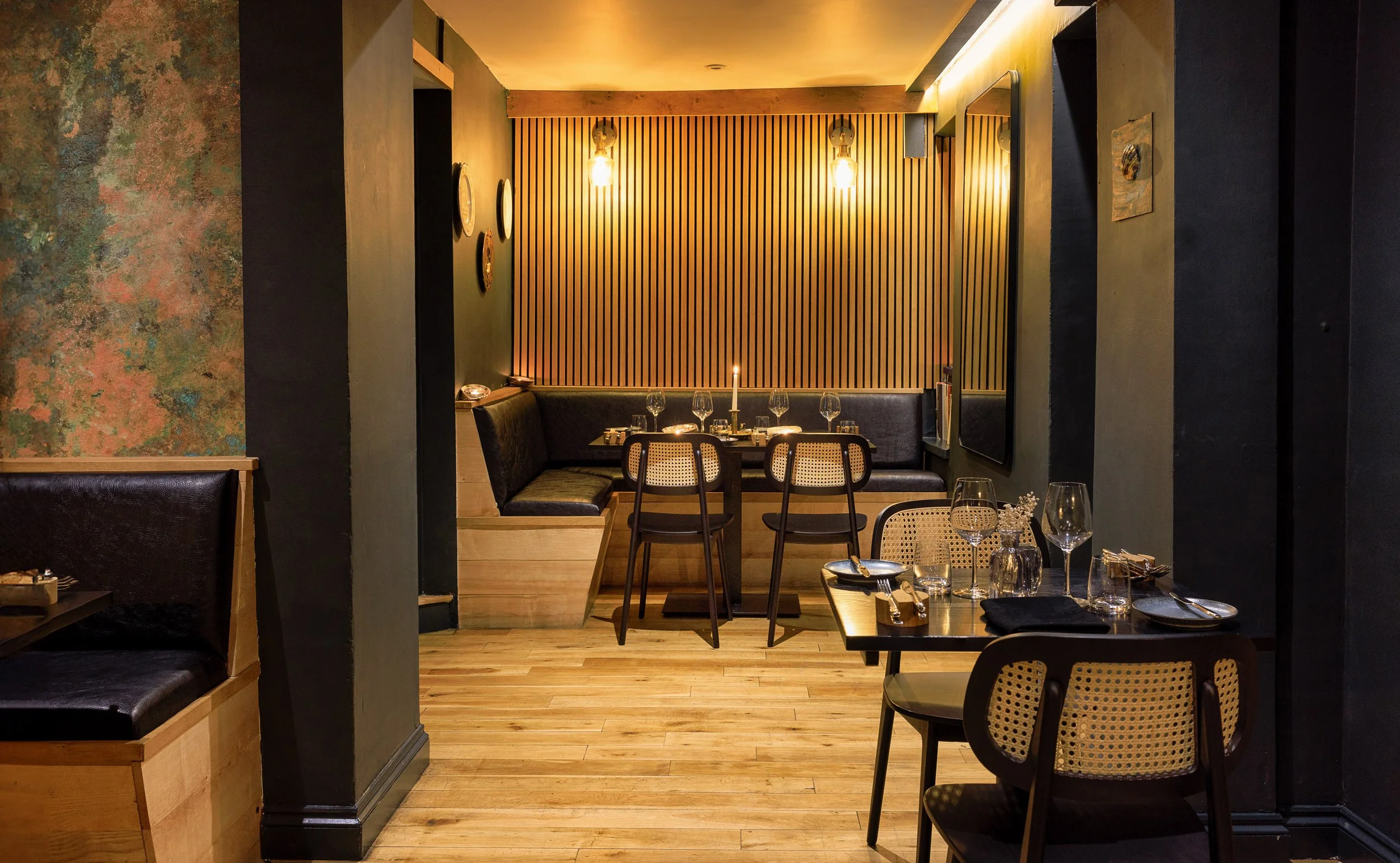 Commercial restaurant interior at Anima Restaurant in Devon, showing dark walls, timber slats, booth seating, cane-backed chairs and neatly arranged dining tables.
