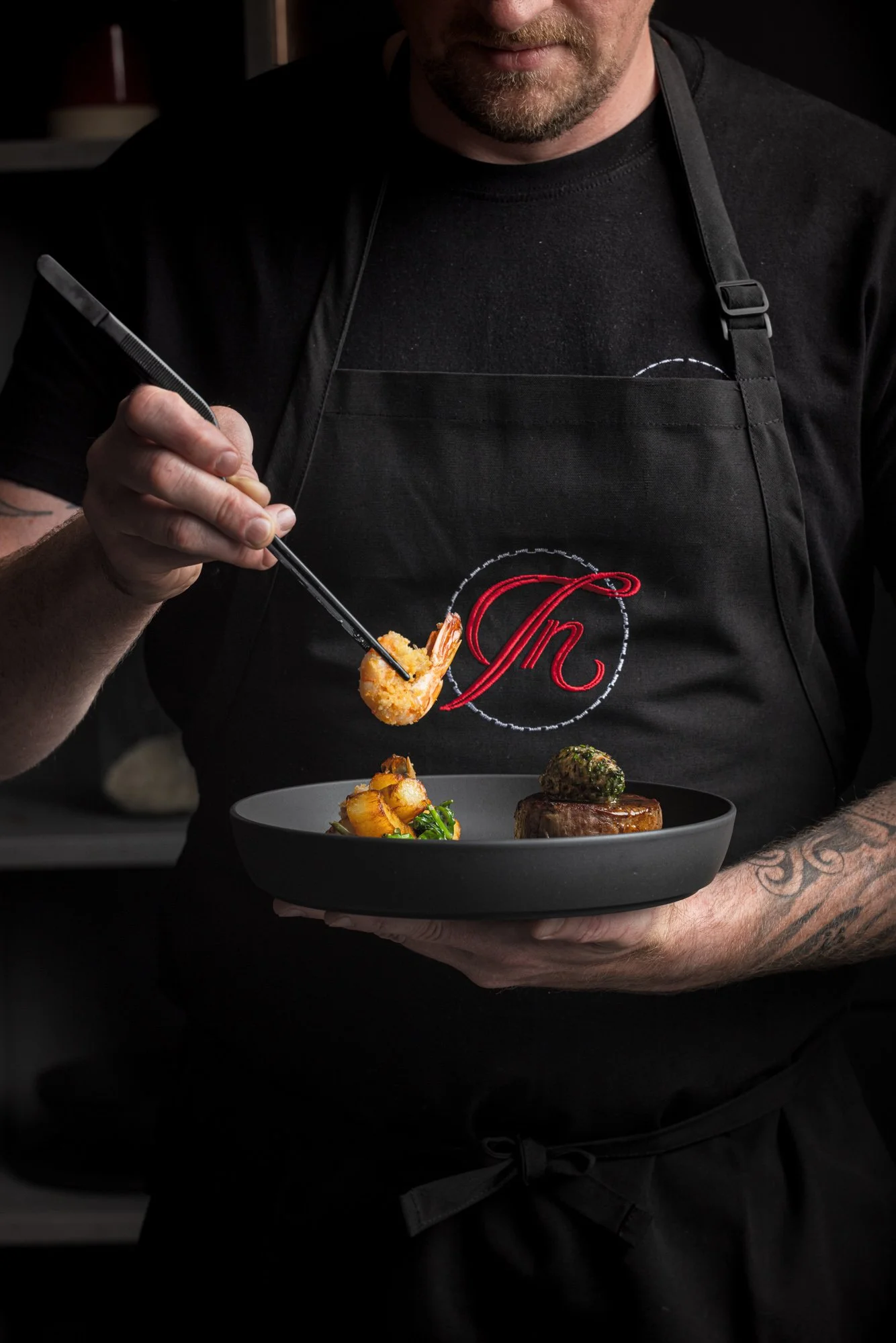 Commercial chef and hospitality photograph of a Dine Indulge chef using tweezers to finish two small dishes while holding a black plate for the Bristol-based private dining company.