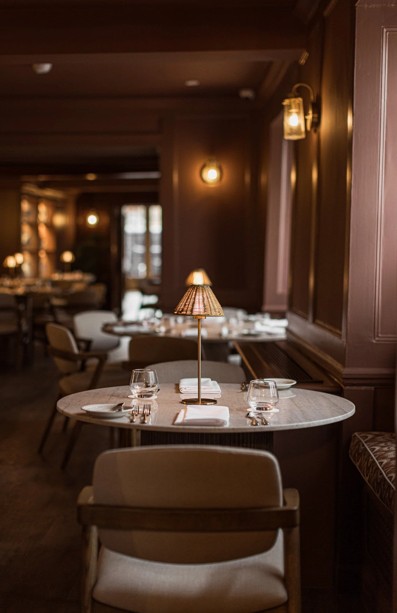 Interior photograph of a neatly set table at The Feathers Hotel in Woodstock, framed by warm panelling, a shaded lamp and softly lit dining spaces beyond.