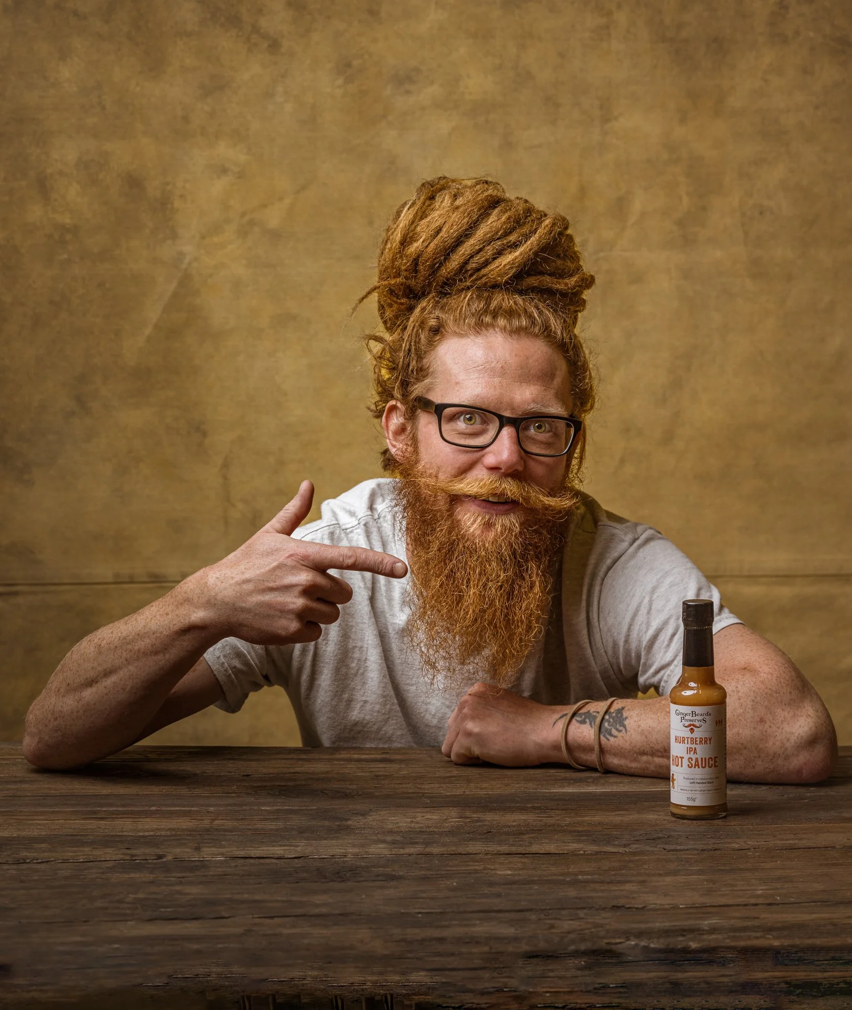 Commercial portrait of a red-haired, bearded maker seated behind his bottled sauce and pointing towards the product, using a warm textured background and relaxed expression for Ginger beard's preserves editorial feature in Bristol.