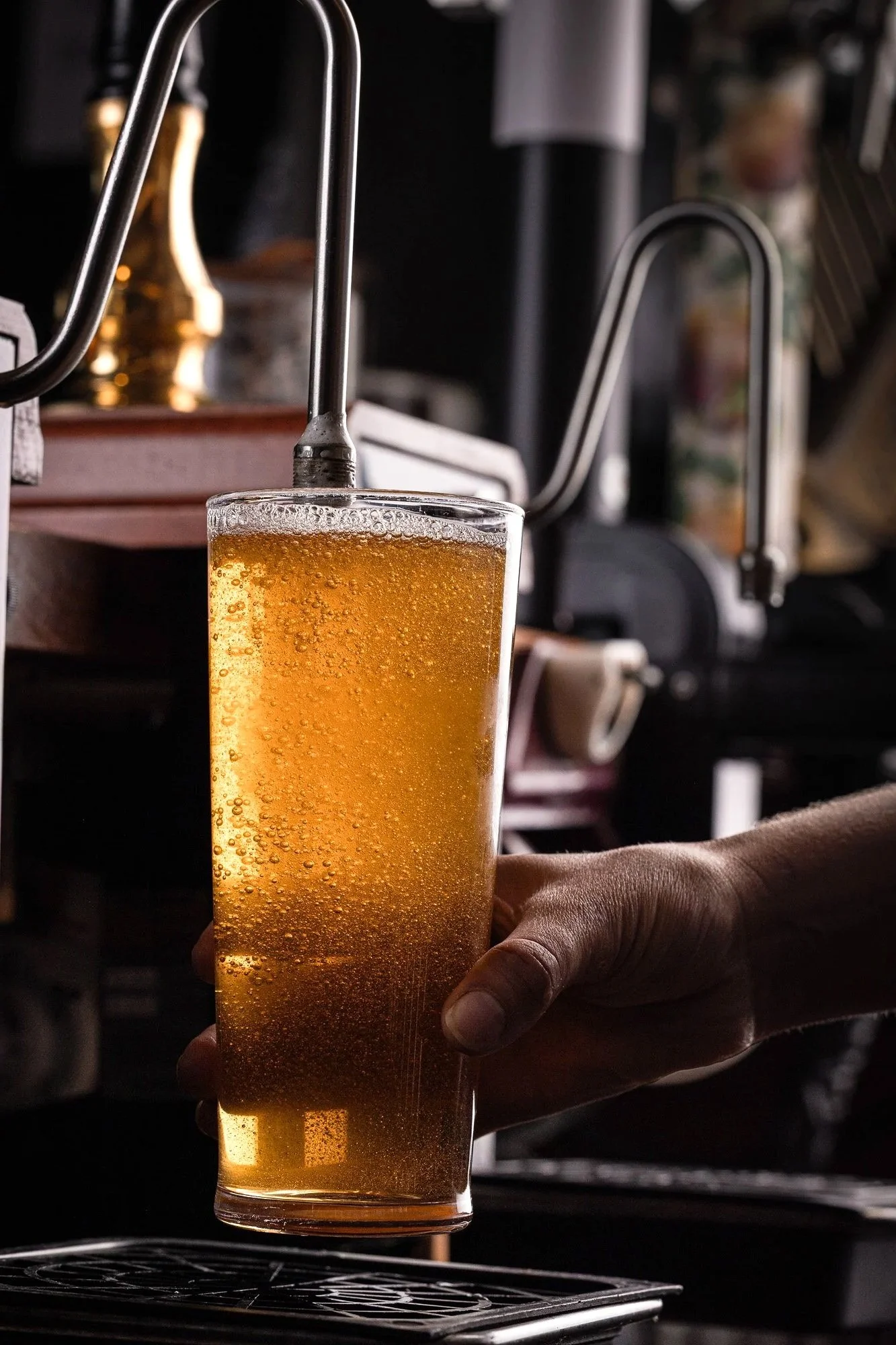 Commercial cider photograph of a hand holding a clear pint of golden cider at a bar, using warm ambient light and visible condensation to communicate refreshment and informal hospitality service.