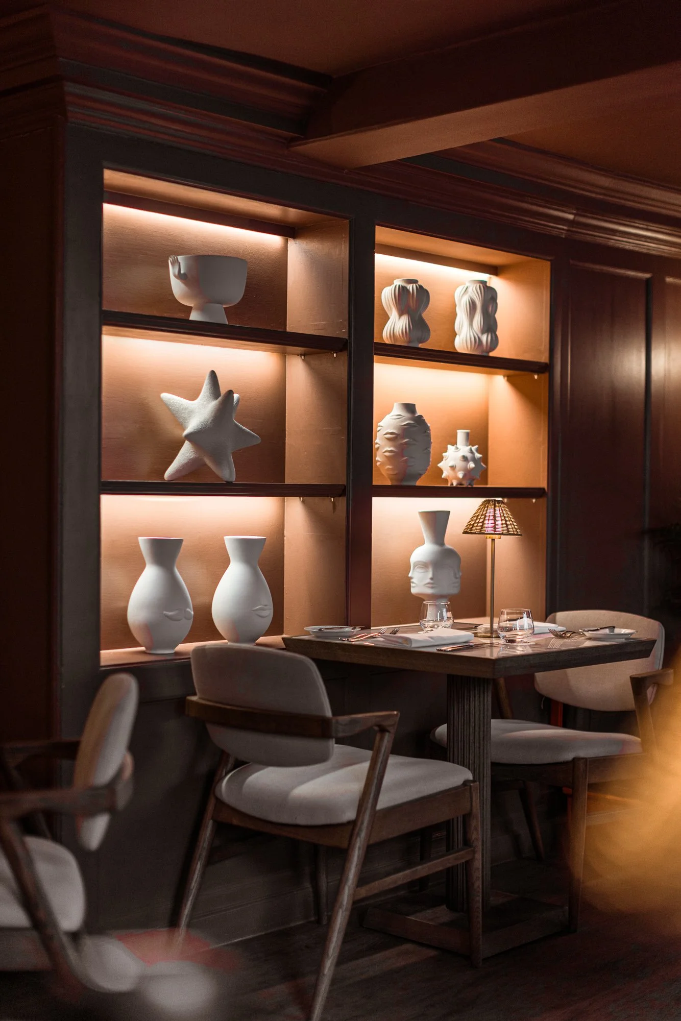Commercial restaurant detail at The Nest Restaurant, The Feathers Hotel, showing illuminated shelves of sculptural ceramics above an intimate dining table.