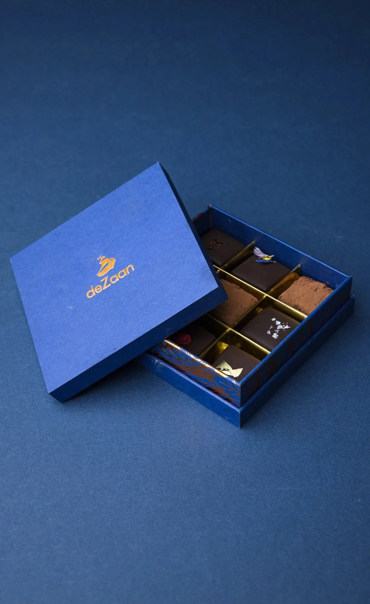 Commercial product photograph of an open blue deZaan presentation box containing assorted chocolates, arranged diagonally on a matching blue background to emphasise the premium packaging and restrained brand design.