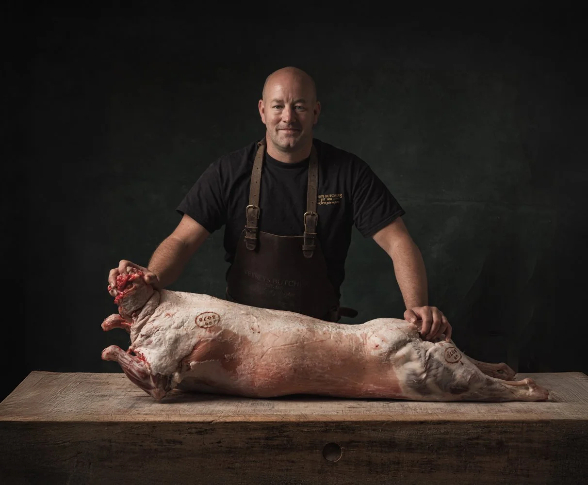Environmental portrait of a butcher standing behind a whole pig carcass on a worktable, combining commercial people photography with the practical setting of a professional butchery in Cornwall.