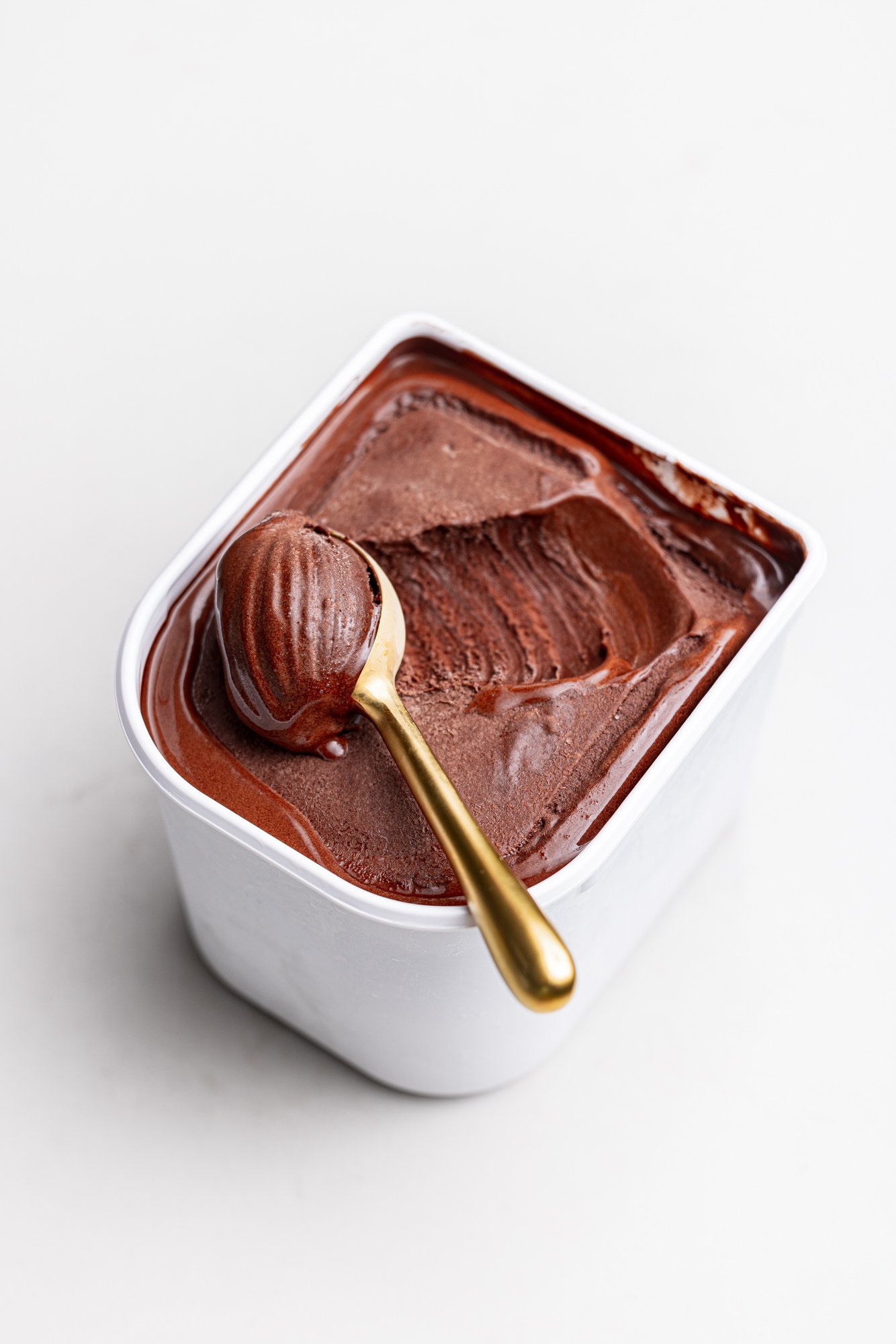 Clean commercial product photograph of an open tub of chocolate ice-cream with a spoon resting across its smooth surface, presented on a pale background for Langage farm packshot website.