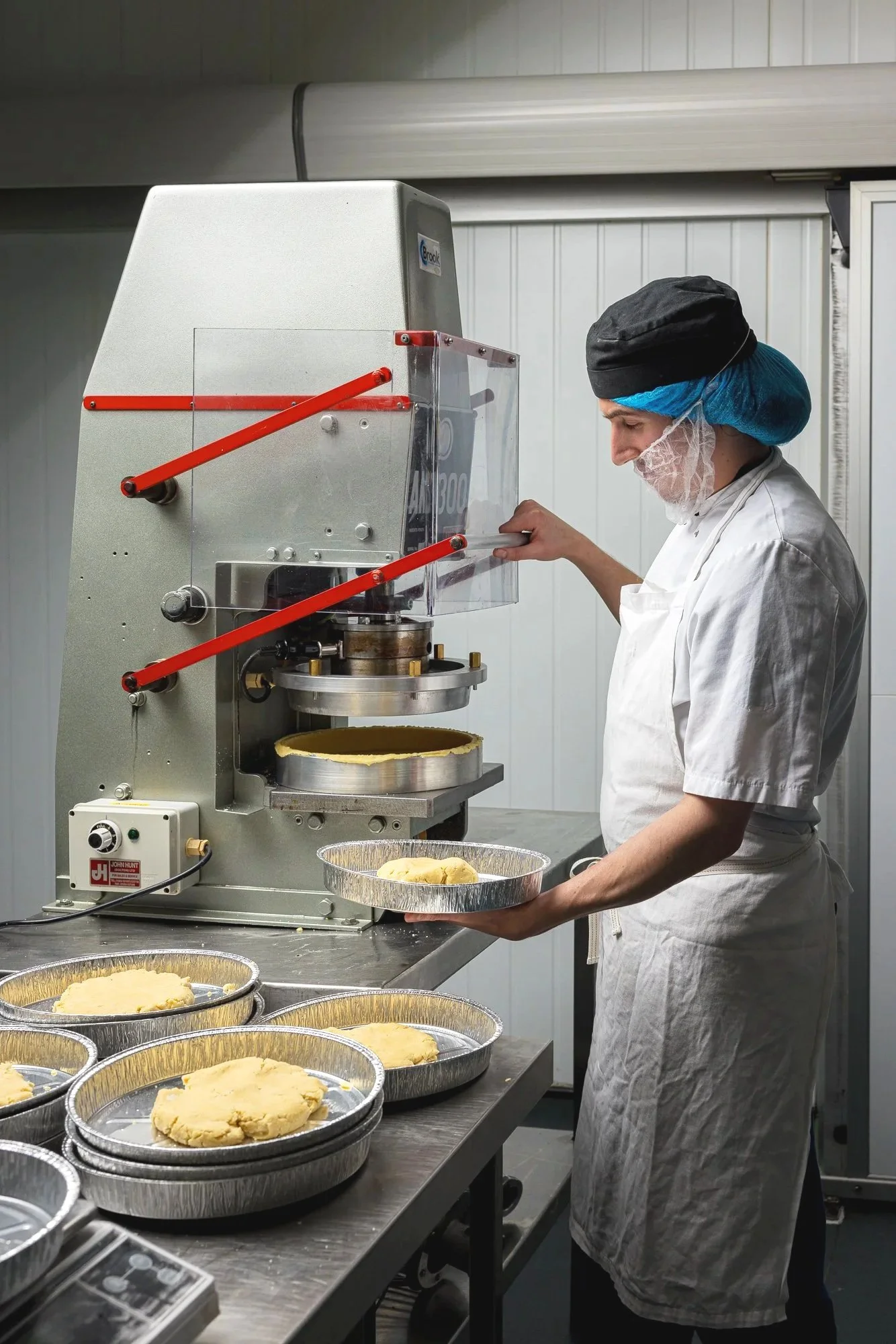 Commercial production photograph of a worker operating specialist equipment beside rows of prepared pies, documenting the person, machinery and practical manufacturing process for Dessertology.