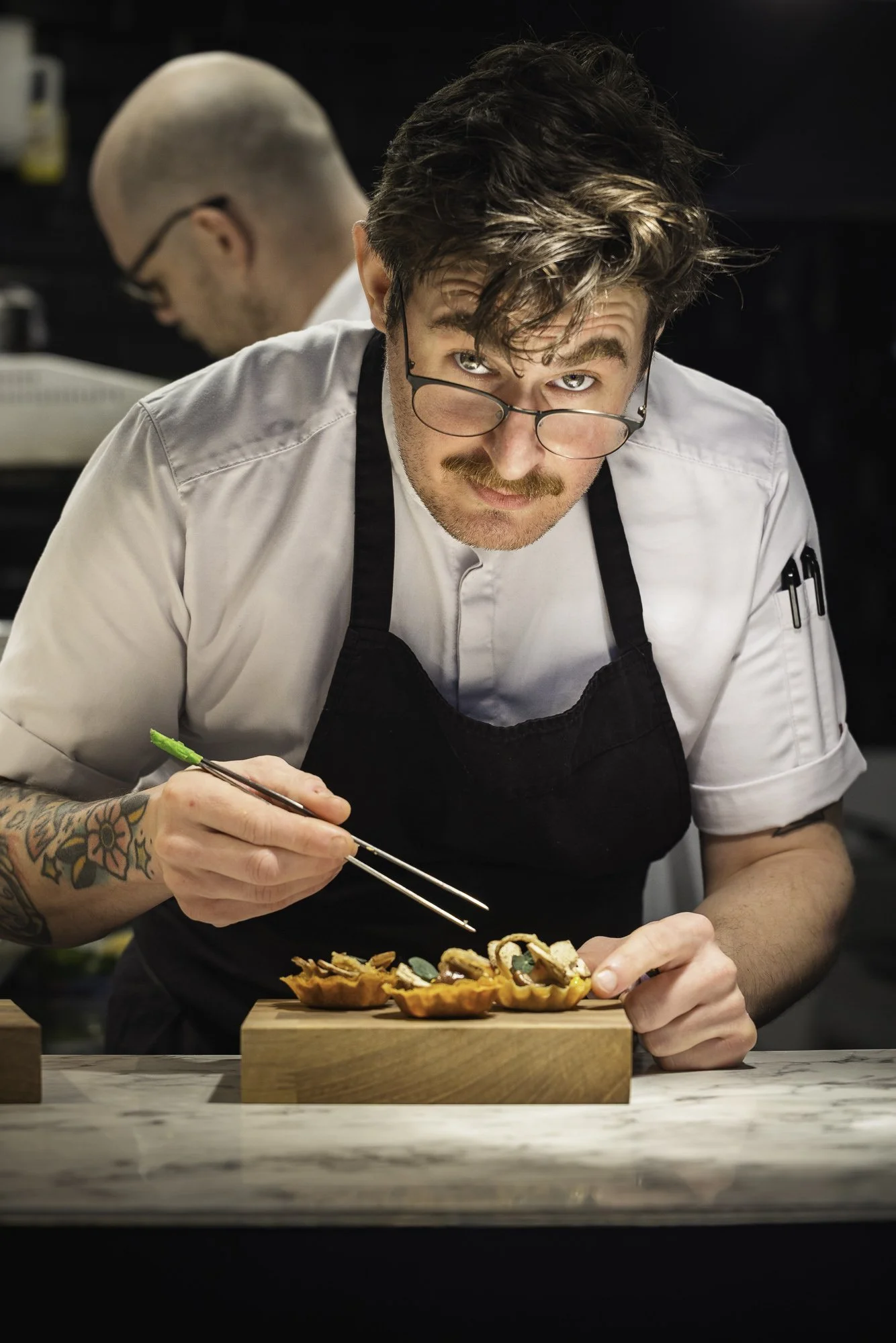 Environmental chef portrait showing a cook leaning closely over a dish while adding the final details with tweezers, using direct expression and the working kitchen setting to communicate precision and concentration.