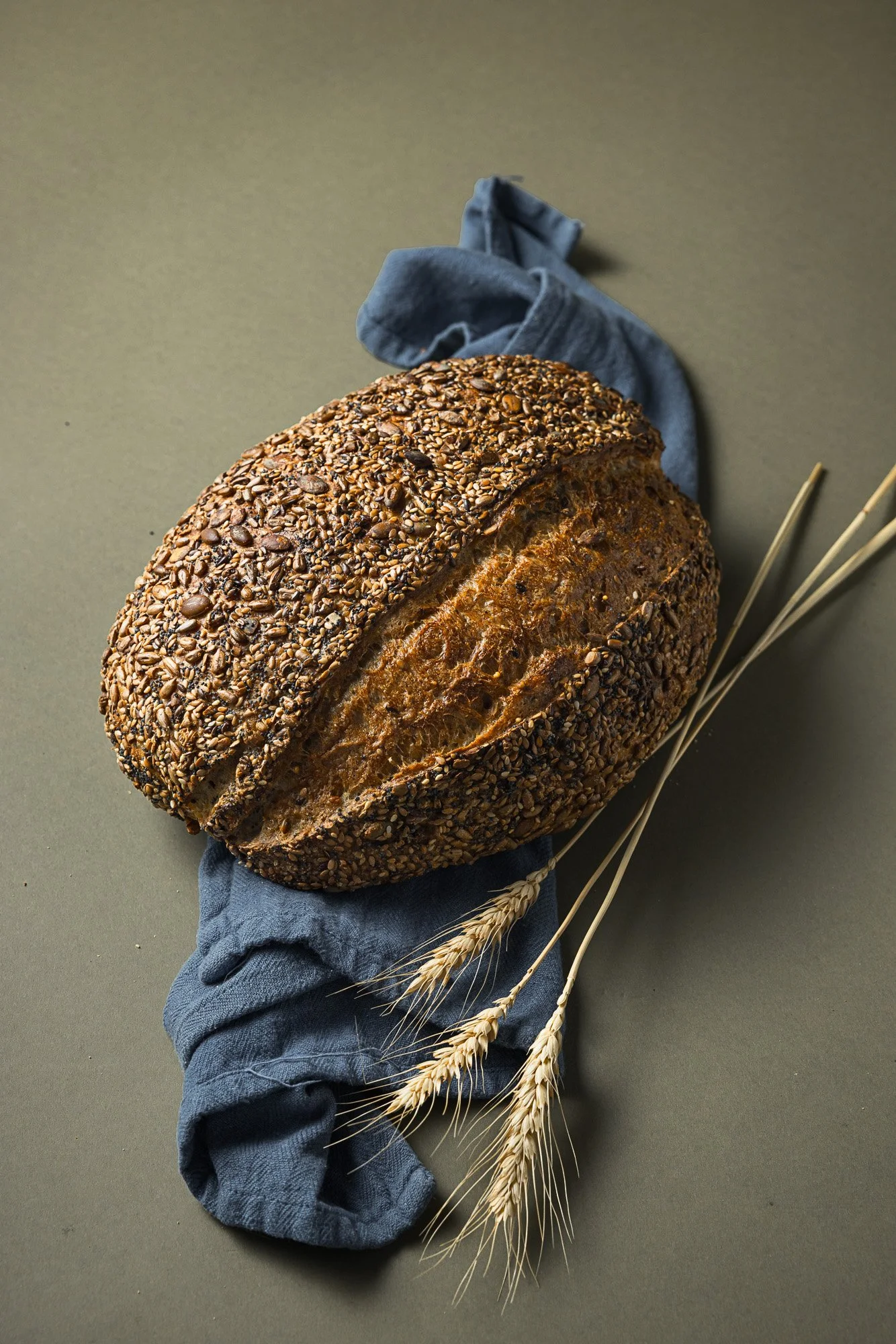 Styled commercial photograph of a seeded artisan loaf resting on folded blue cloth with wheat stems, using a muted background and soft directional light to emphasise crust and seed texture.