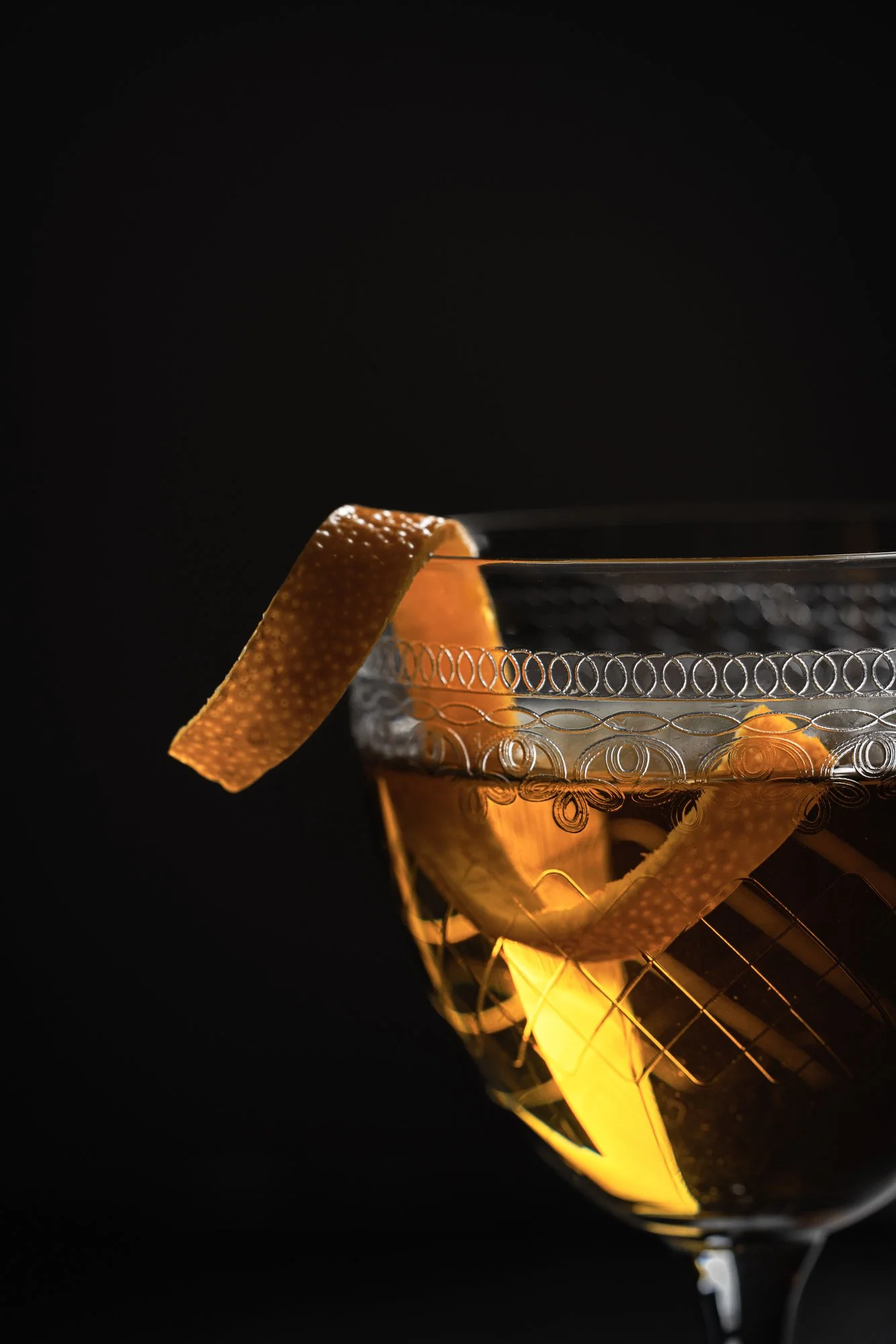 Close commercial photograph of an amber cocktail served in a cut-glass coupe with a broad orange peel garnish, using dark directional lighting to emphasise the drink’s warm colour, glass detail and citrus oils.