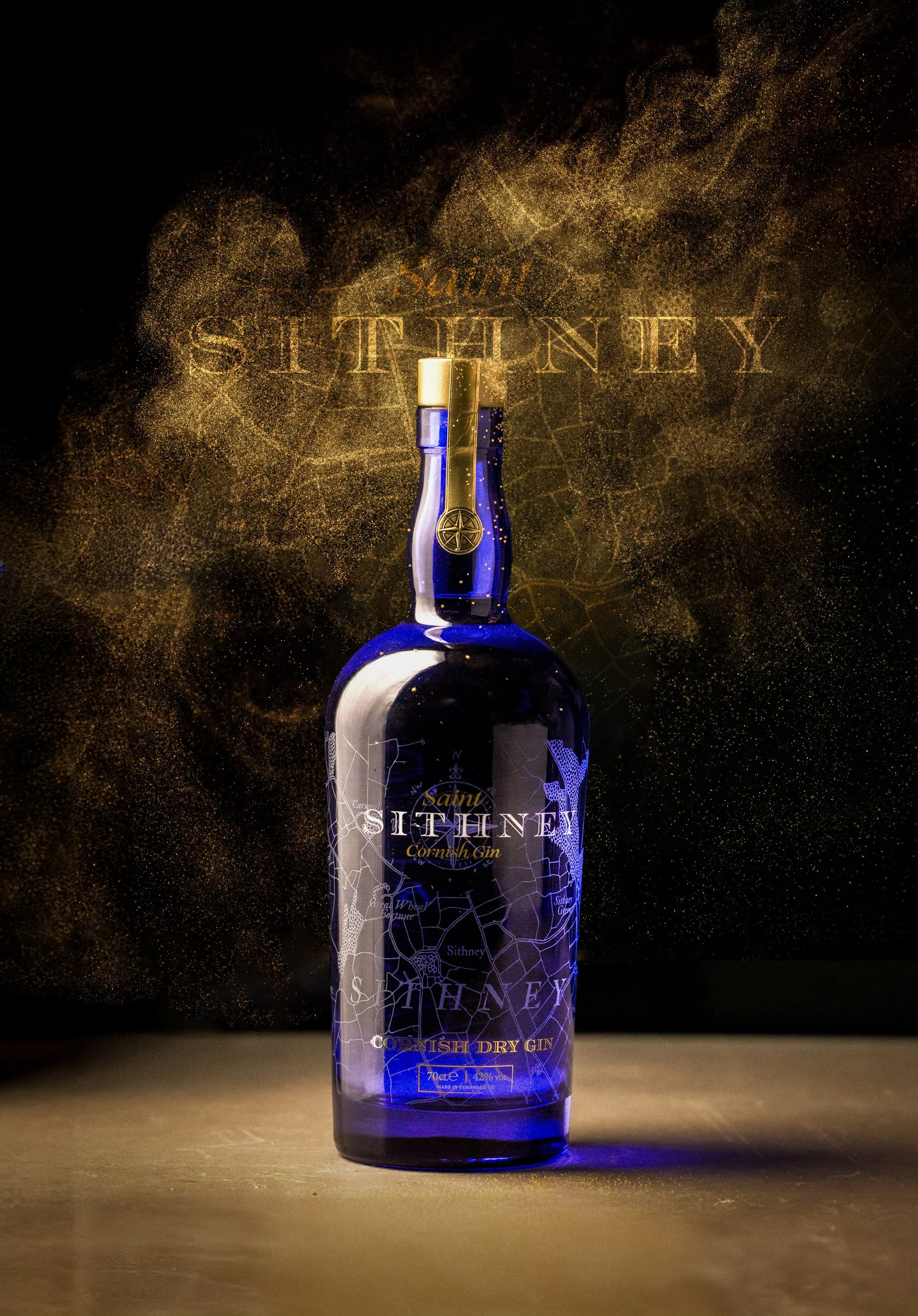 Dramatic commercial bottle photograph of a deep blue Saint Sithney surrounded by suspended golden smoke against a black background, using controlled studio lighting to reveal the glass colour, label and silhouette.