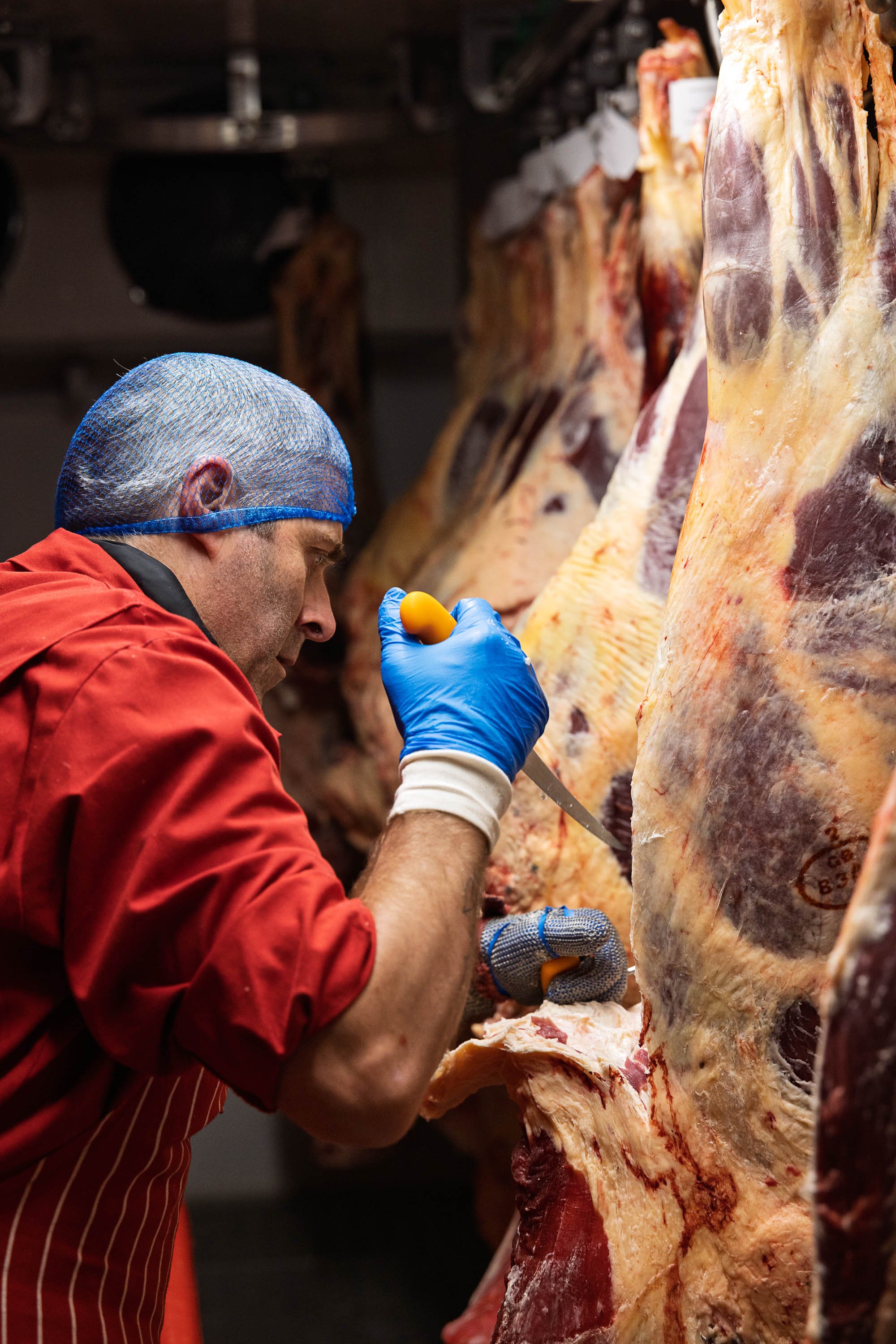 Documentary-style butchery photograph of a butcher in protective clothing working closely on a hanging beef carcass, recording the production environment and skilled preparation for Veyseys Butcher in Devon.