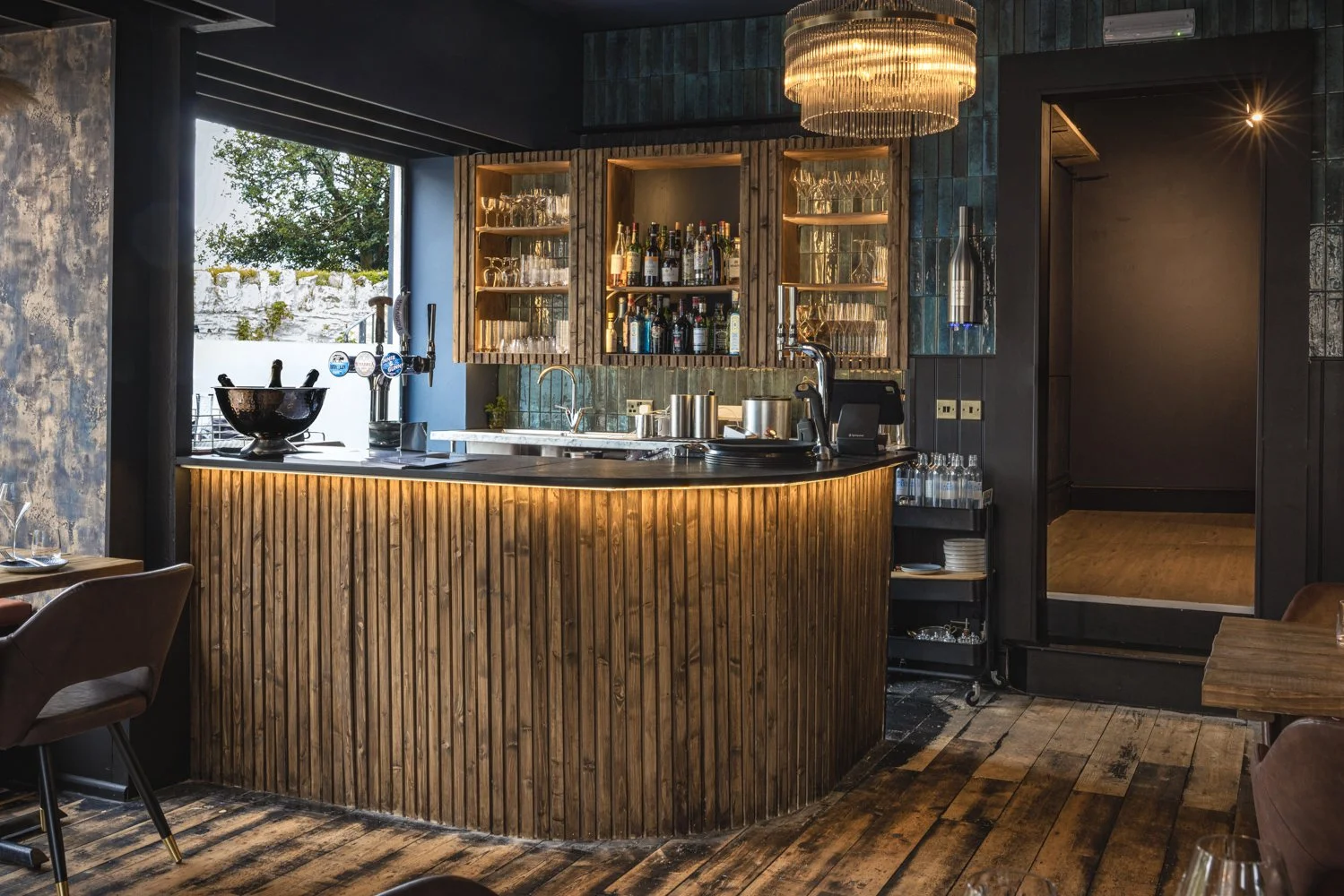 Commercial bar interior at Kensington Restaurant in Lynton, Devon, showing a timber-fronted counter, illuminated bottle cabinets, glassware, beer taps and a statement ceiling light.