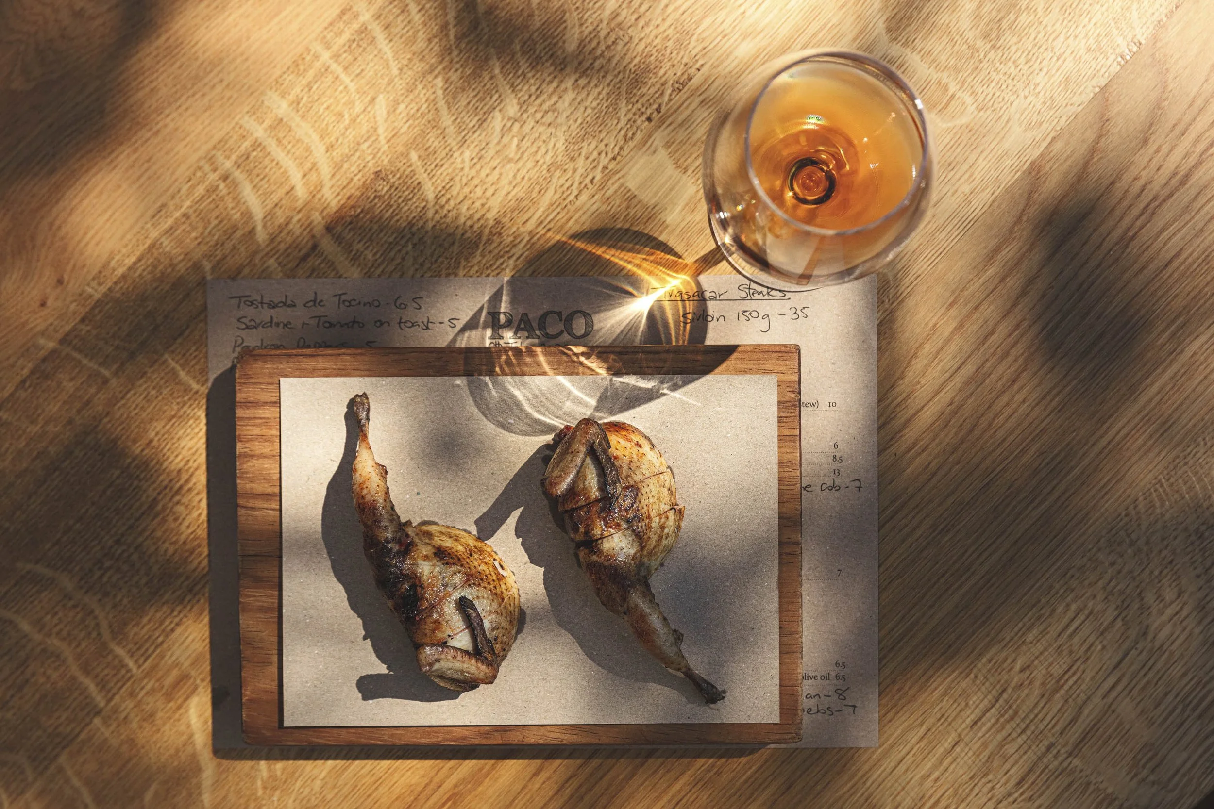Editorial restaurant photograph of two roasted pigeon presented on a wooden board with wine, handwritten kitchen notes and strong afternoon shadows for Michelin starred Paco Tapas in Bristol.