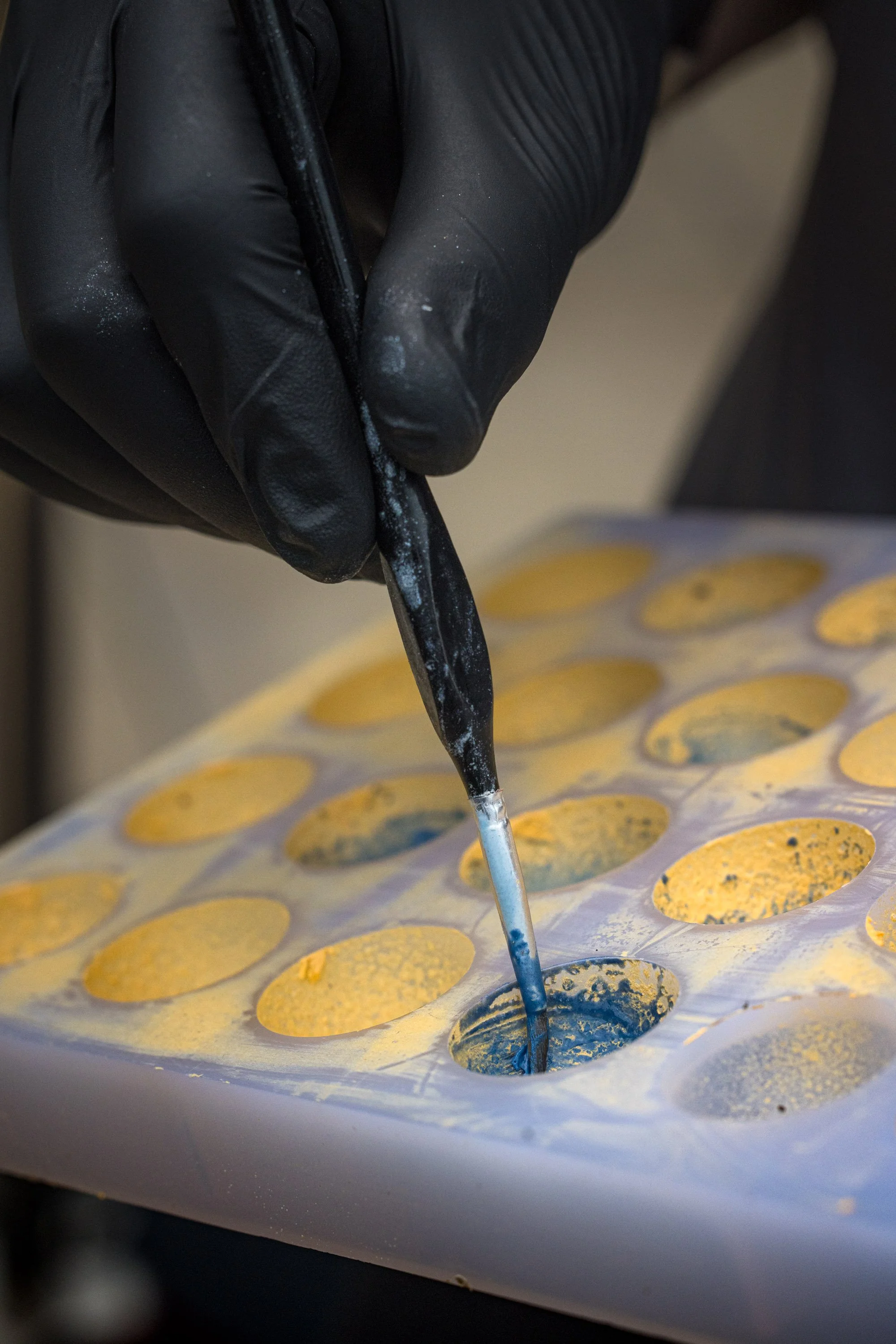 Commercial production photograph of a gloved hand filling rows of chocolate moulds with coloured mixture, documenting the precise preparation process for chocolatier, bakery or food manufacturer in location