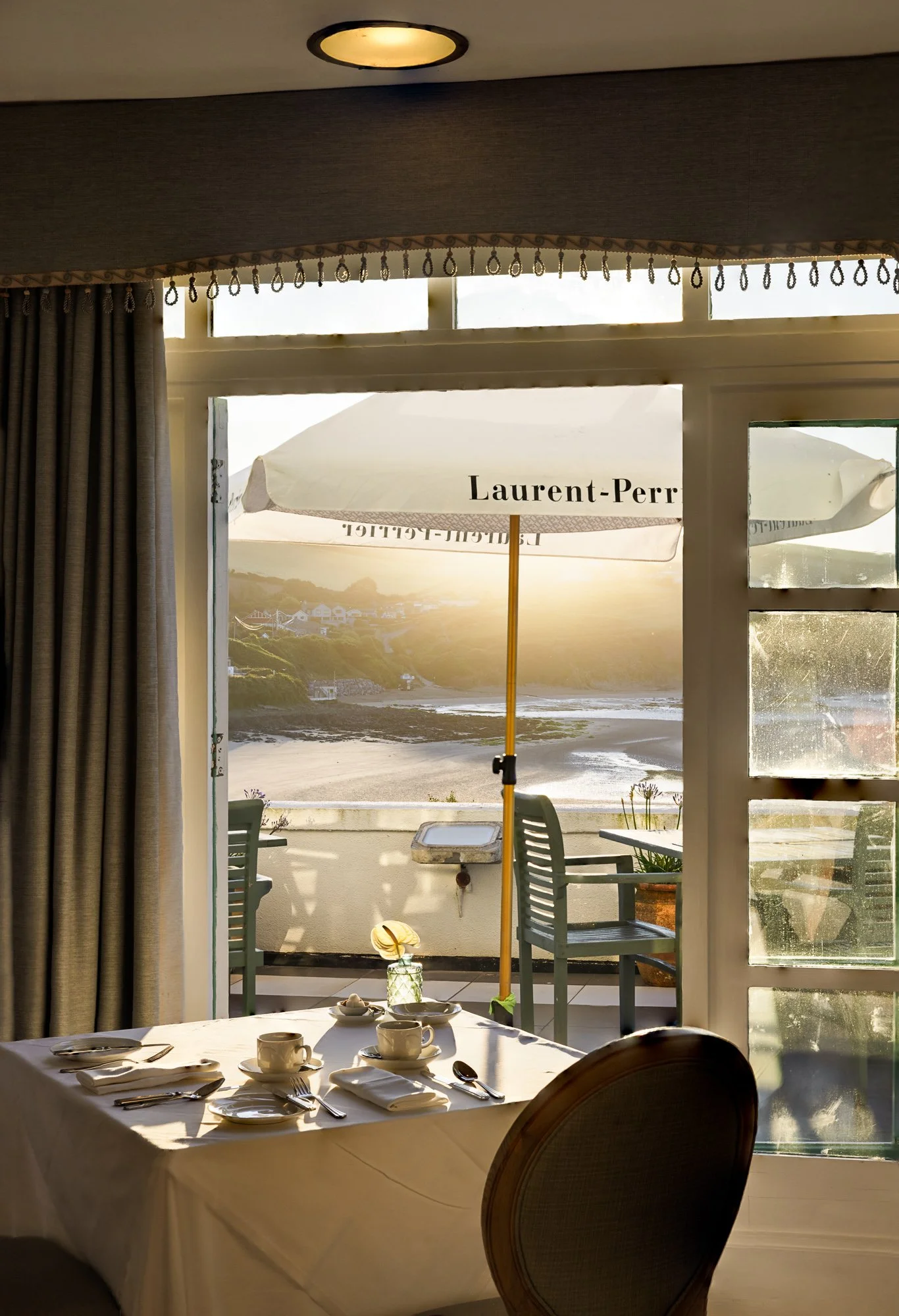Commercial hospitality interior at Burgh Island Hotel in Devon, showing a laid table beside large coastal windows with the terrace, sea view and Laurent-Perrier umbrella outside.