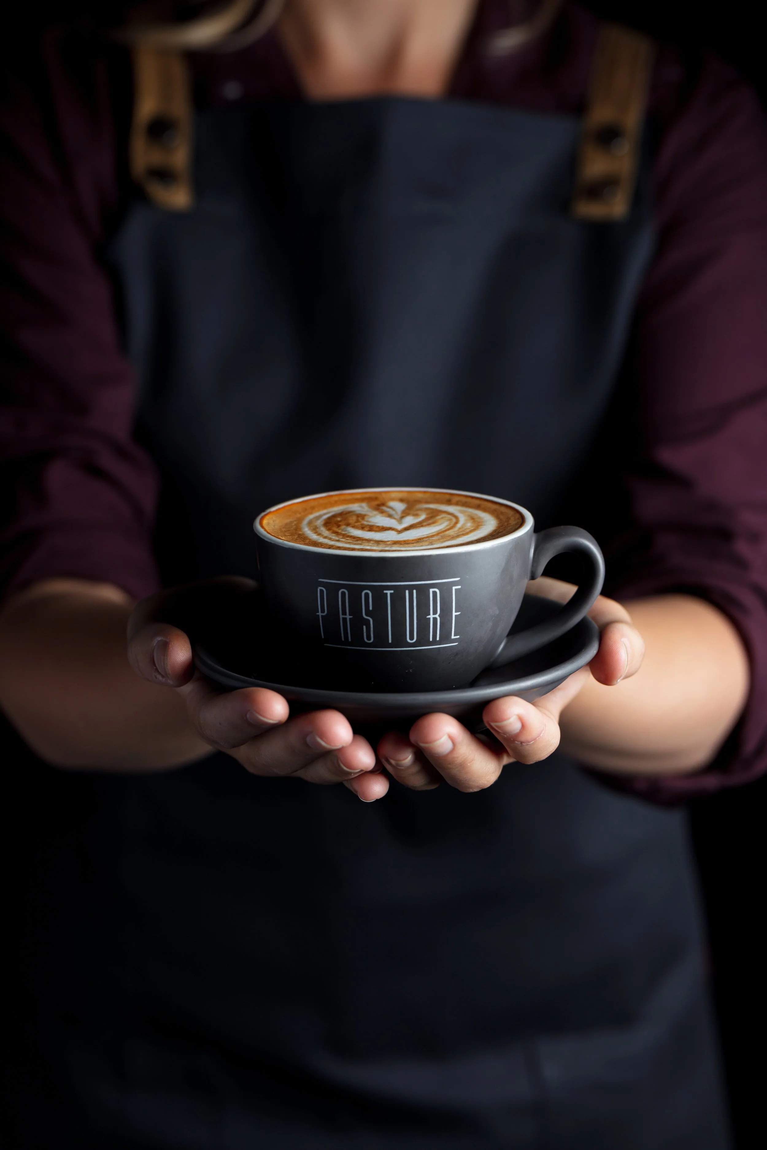 Commercial coffee photograph of a Pasture-branded cup and saucer held carefully in two hands, with detailed latte art visible against a dark background to emphasise warmth, service and presentation.