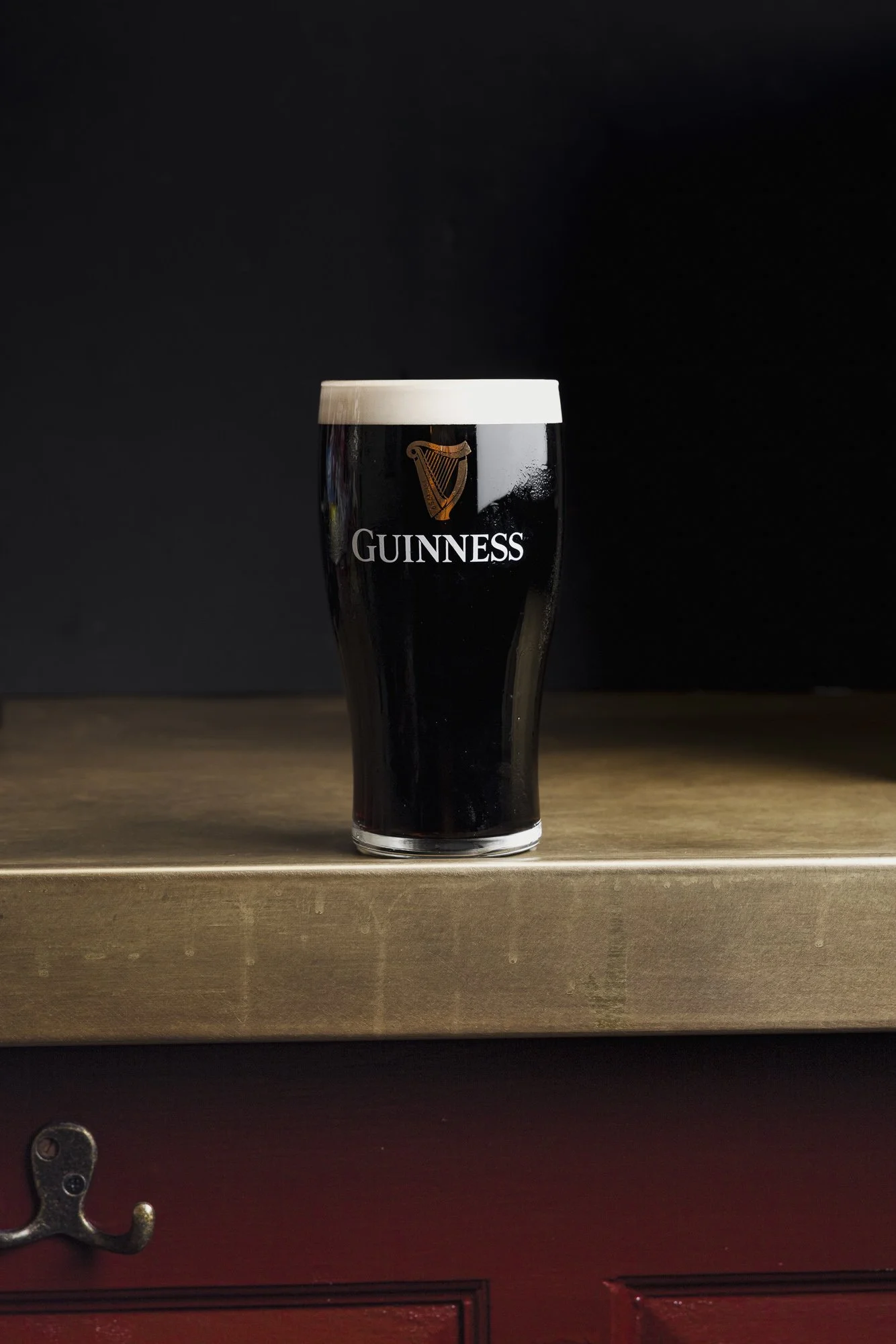 Minimal commercial beer photograph of a freshly poured pint of Guinness with a clean white head, presented against a near-black background on a warm bar surface to emphasise the iconic glass and stout colour.