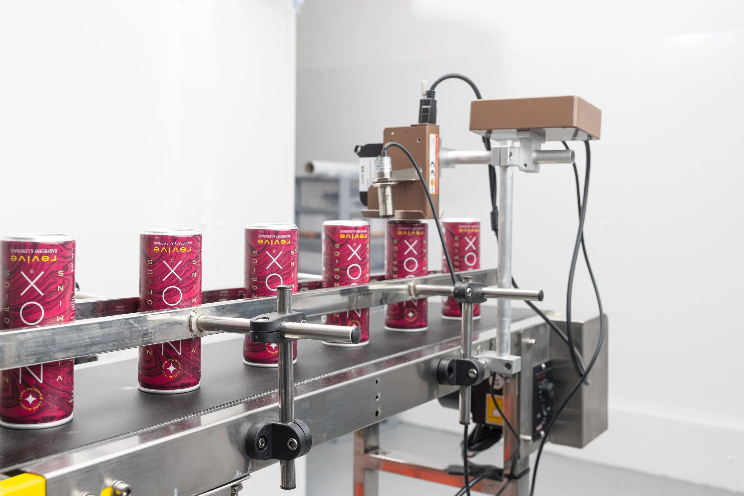 Commercial manufacturing photograph of branded drinks cans moving through a compact conveyor system, documenting the packaging stage and production equipment used by beverage producer in London.
