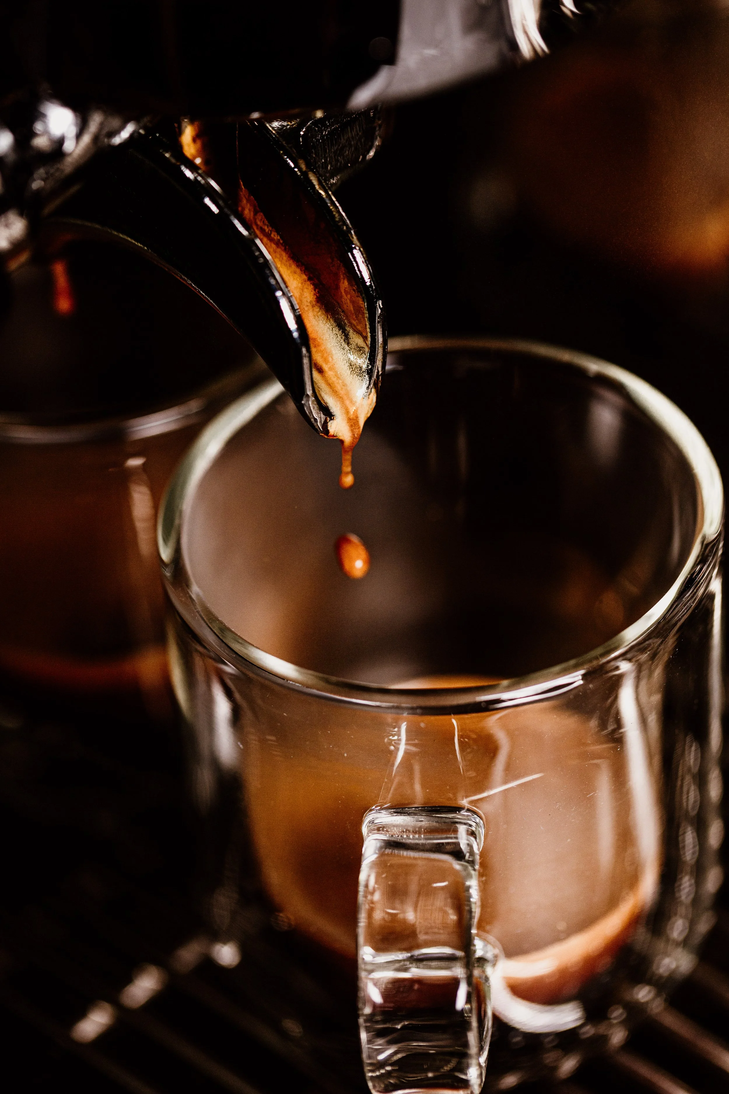 Macro commercial drink photograph of espresso coffee pouring from coffee machine into a clear glass cup, capturing the suspended droplet, reflections and warm colour at the moment of pouring.