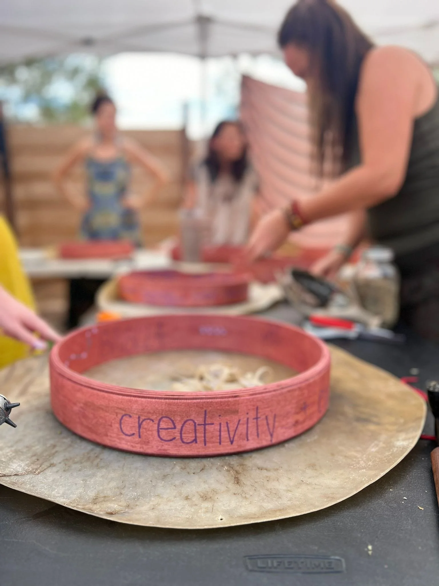 Pink wristband with the word "creativity" written on it, resting on a stone surface, with a blurred background of people working and talking under a canopy.