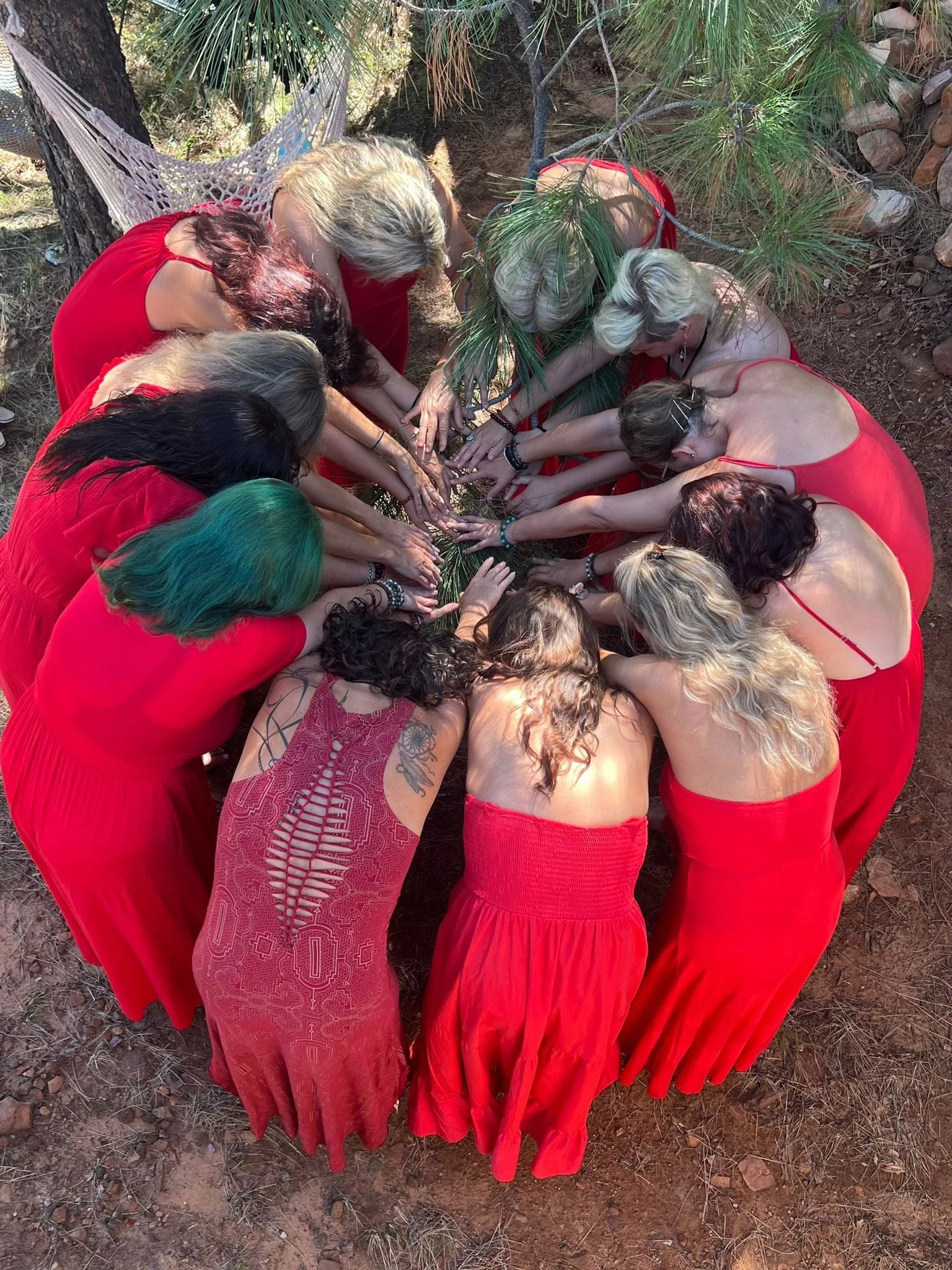A group of women dressed in red, with some wearing sleeveless dresses, are gathered in a circle outdoors. They are placing their hands together over a green pine branch, engaged in a collective activity.
