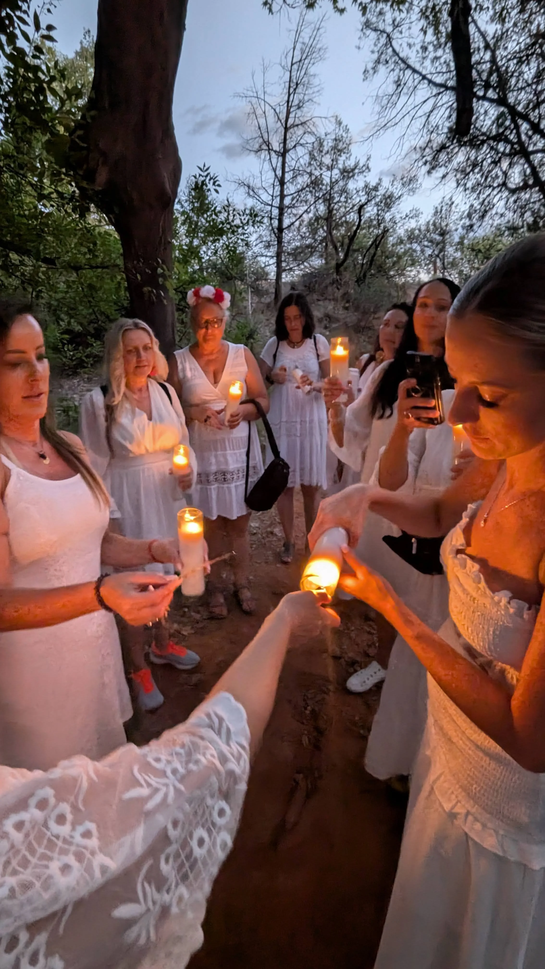 A group of women in white dresses holding candles, gathered outdoors in a wooded area in Sedona at dusk, participating in a Slavic Sisters Retreat.