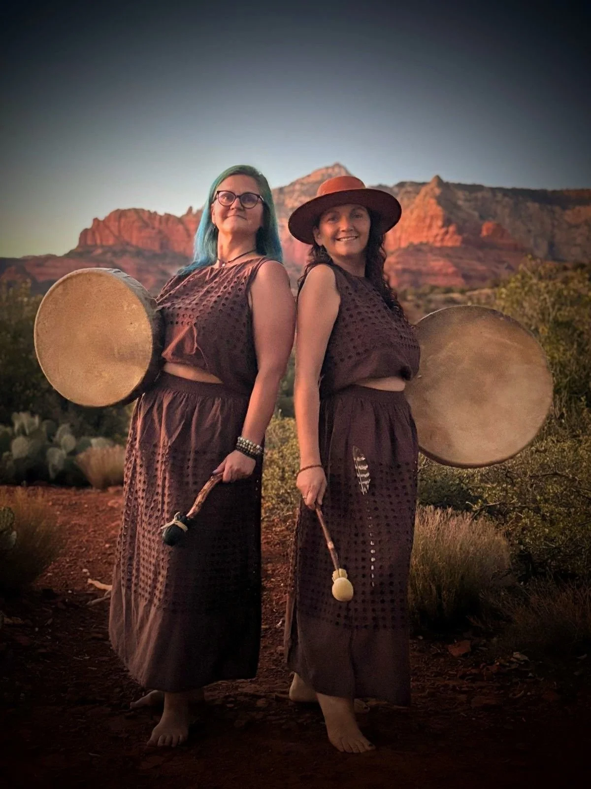 Two women, Slavic sisters, standing outdoors in Sedona with desert landscape, red rock formations, and blue sky in the background, holding large round drums and sticks, dressed in similar brown clothing.