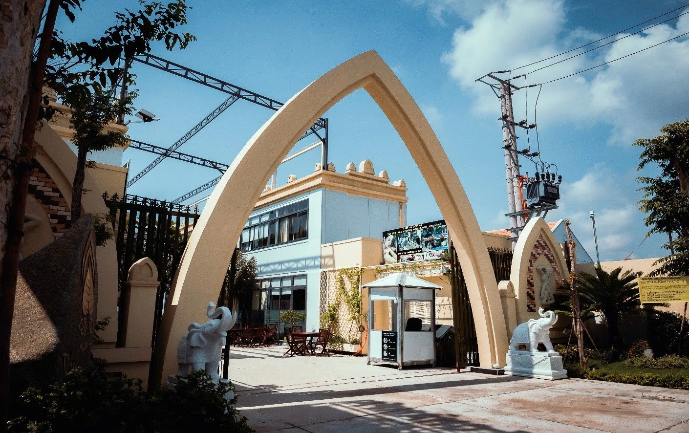 Entrance gate with large decorative archways, elephant sculptures, and a guard post leading into a building complex under a blue sky with clouds.