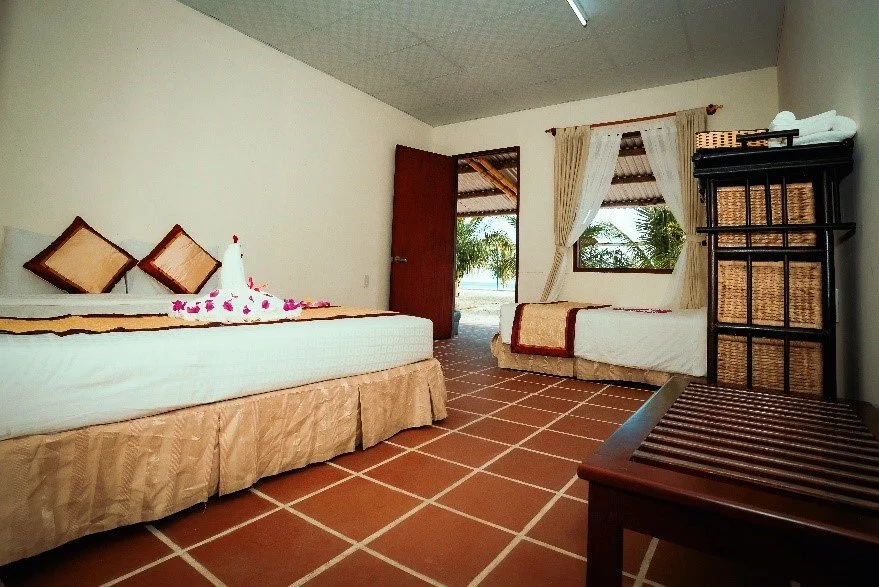 Hotel room with two beds, floral decoration, and a view of trees outside through open door and window.