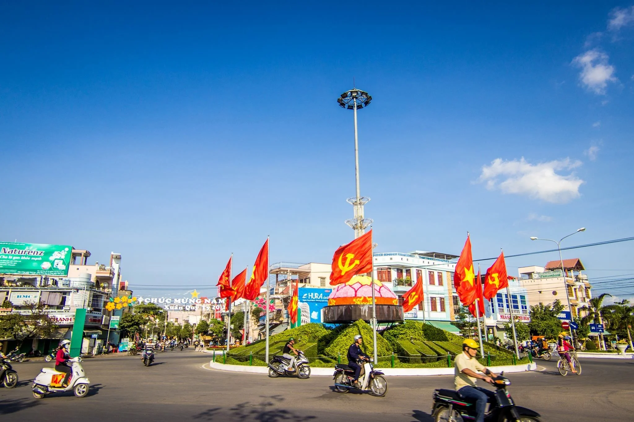 A roundabout with a central monument decorated with red flags featuring a yellow star and hammer and sickle symbols, surrounded by vehicles including motorbikes and bicycles, in an urban setting with buildings and a clear blue sky.