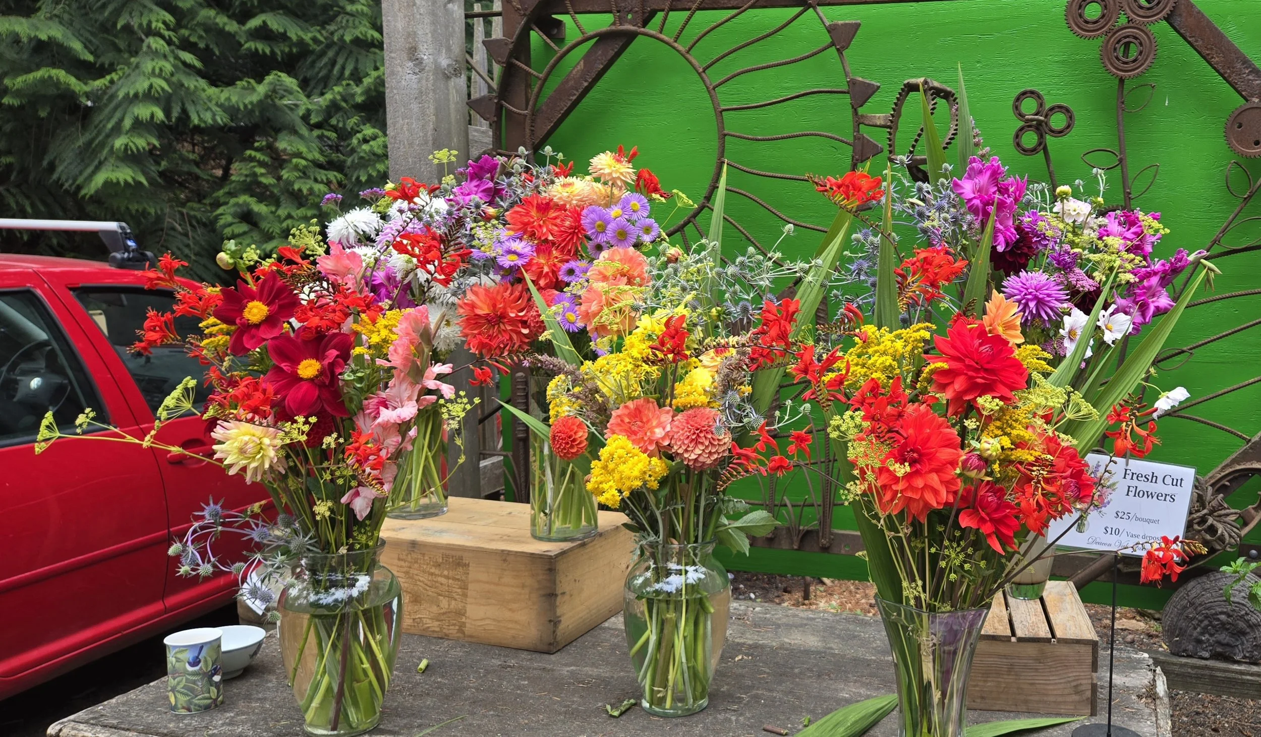 Display of colorful fresh cut flowers in glass vases on a wooden table at a market, with a green background and a traffic light sculpture.