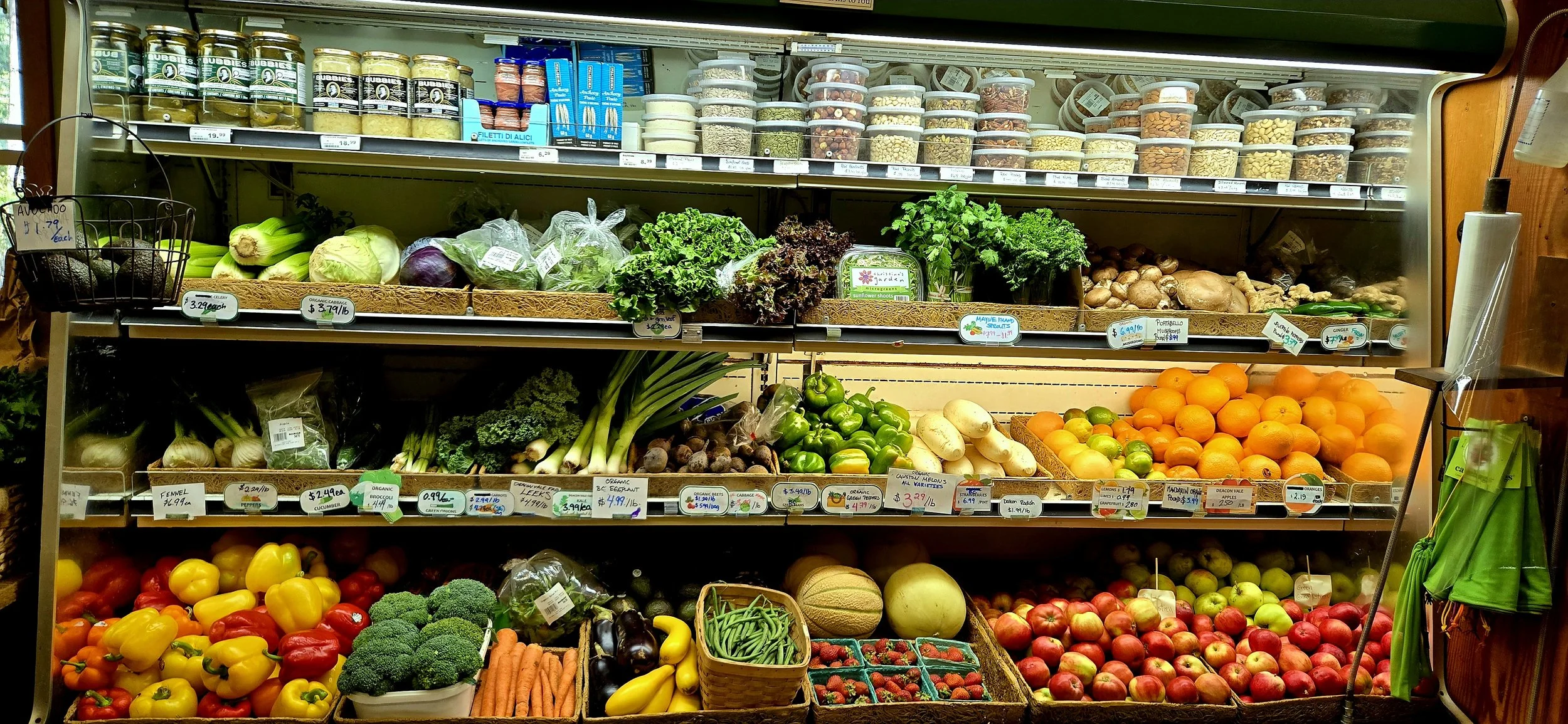 Display of fresh vegetables and fruits in a grocery store, including peppers, broccoli, carrots, leeks, apples, oranges, and other produce.