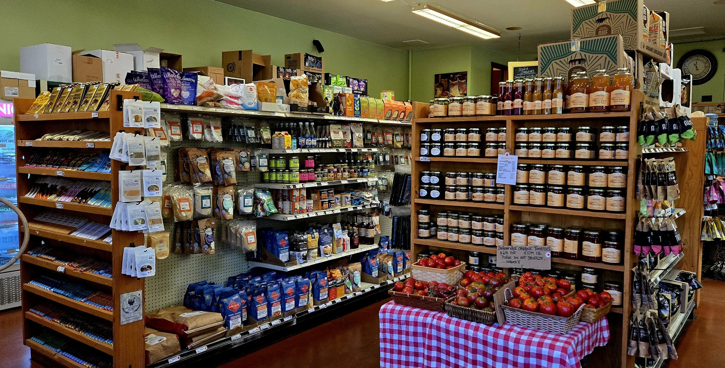 Shelf display of canned goods, jars, and packaged food items, with a basket of fresh tomatoes on a red and white checked tablecloth in front of the shelves, in a grocery store.