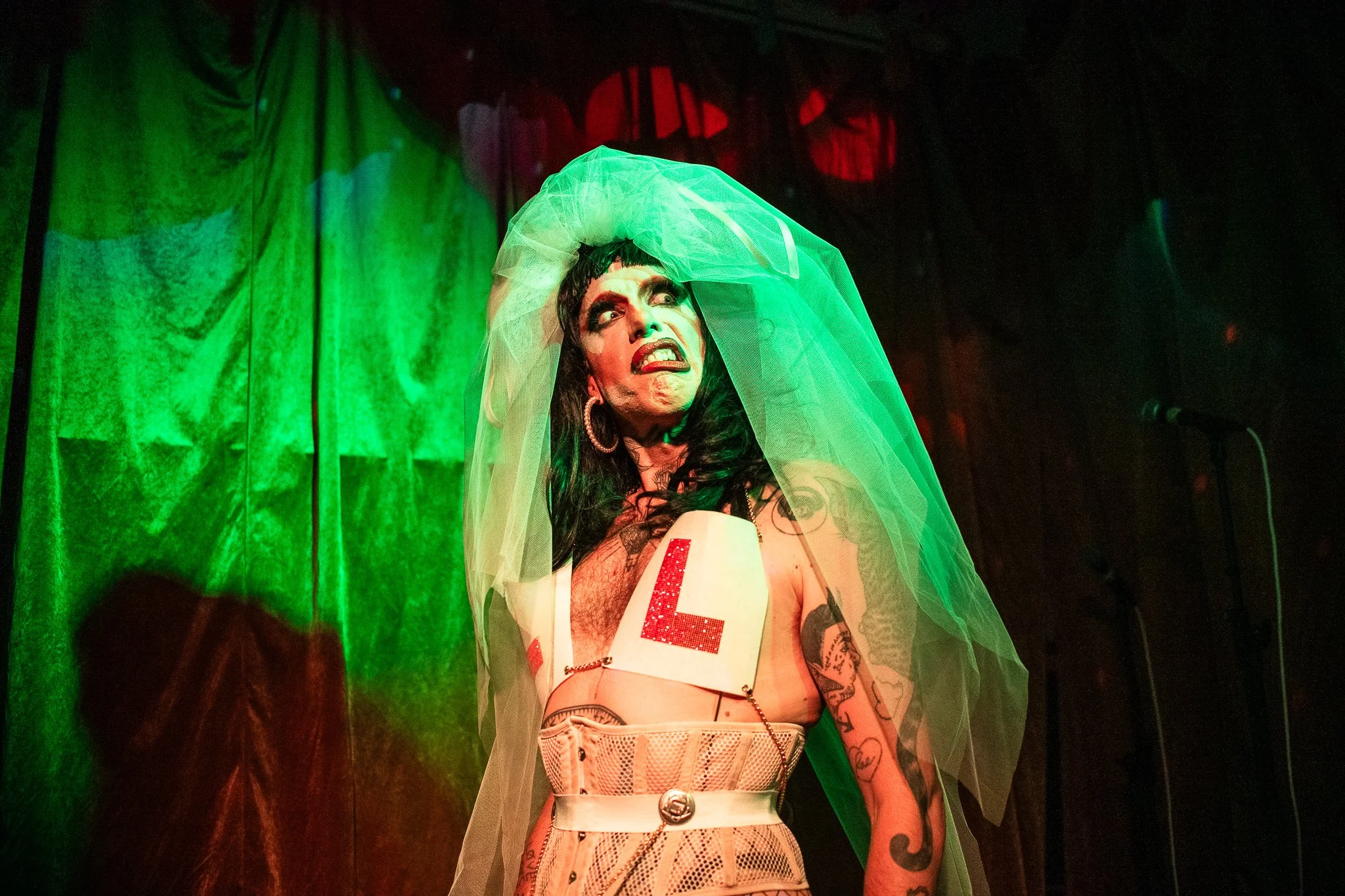 Performer in gothic-themed costume with veil on stage, green lighting, tattoos visible, microphone in background.