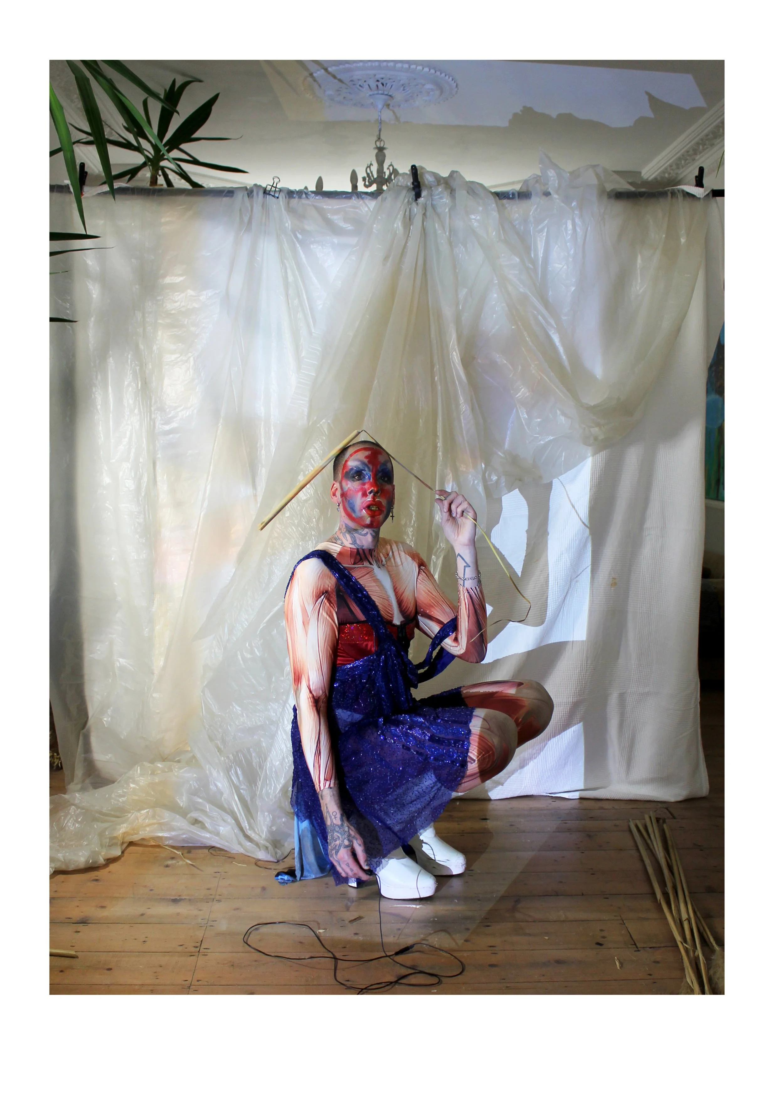 Person in artistic makeup and outfit crouching in front of a backdrop made of draped plastic sheets, with plants and wooden sticks nearby.