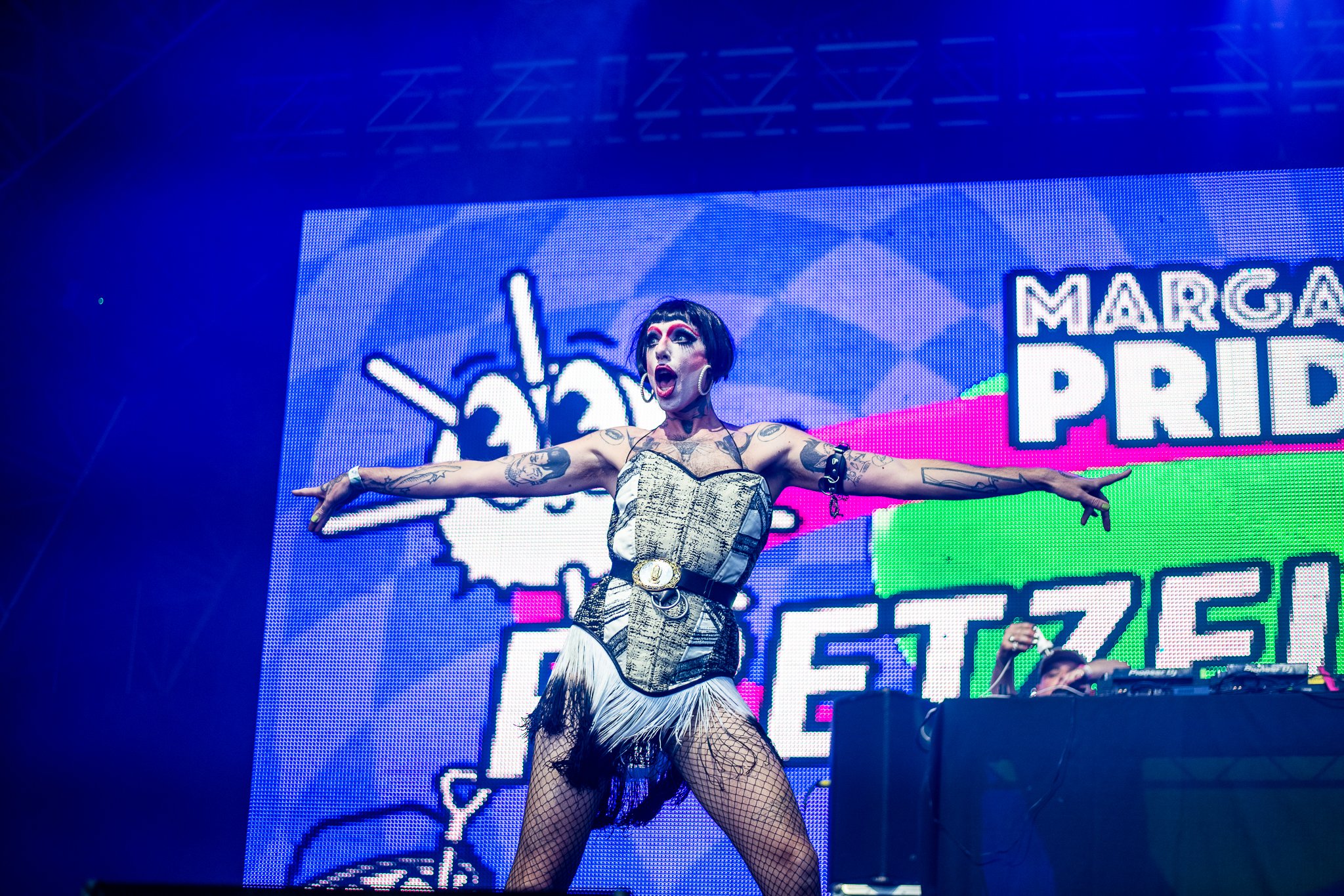 A performer in a flamboyant outfit poses with outstretched arms on stage, with a colorful LED screen background featuring playful graphics and bold text.