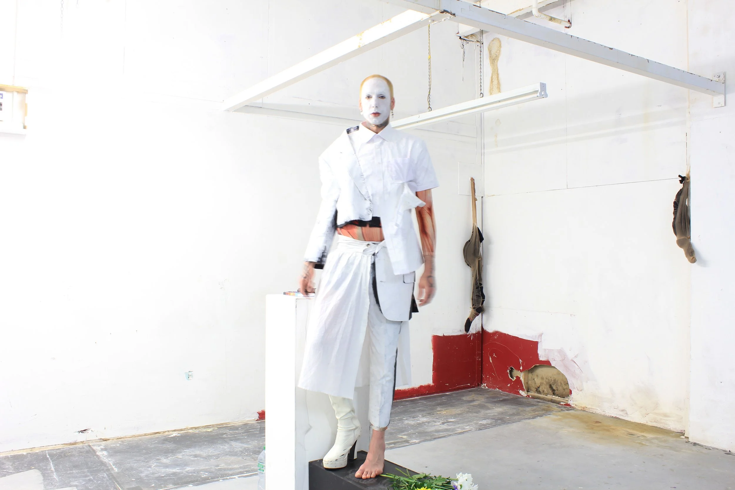 A person in a white outfit and face paint standing in an art studio, with one shoe on and one barefoot, surrounded by industrial-looking decor and objects on the wall.