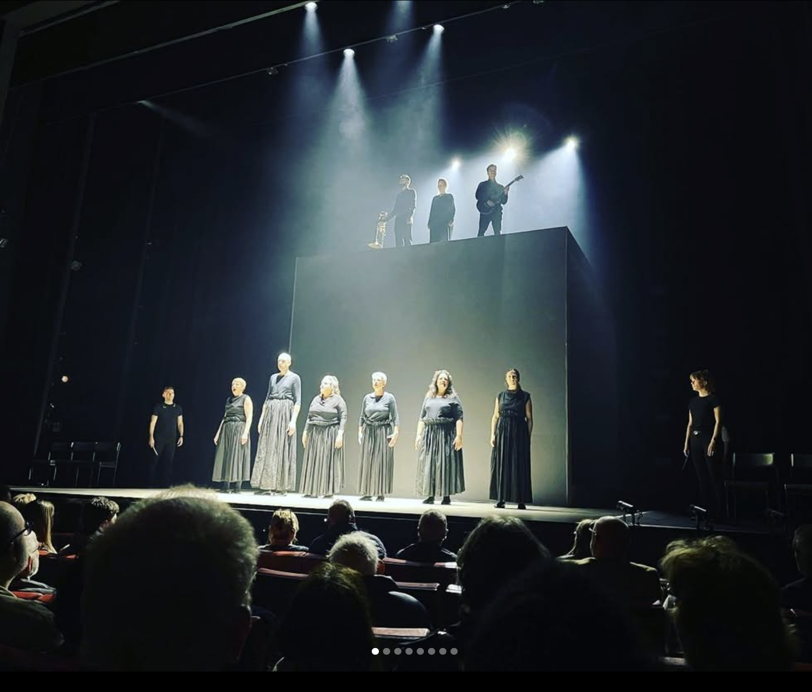 Theater stage with seven women standing in formation, dressed in long skirts and black tops, performing a scene or song. Three people are on an elevated platform above them, with two women and a man playing guitar and standing, under dramatic stage lighting.