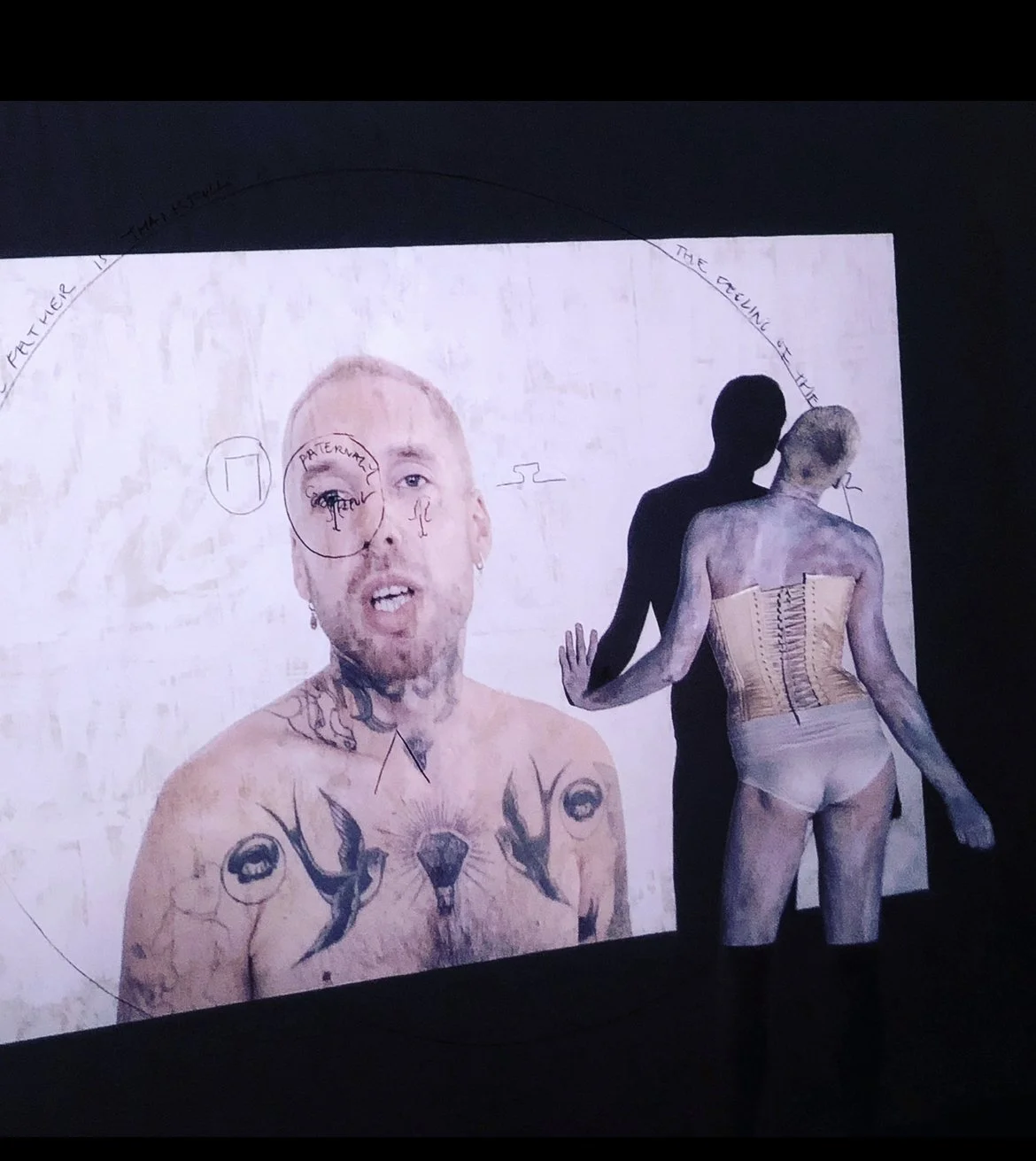 Person in a corset touching a wall with art projection featuring a tattooed individual.