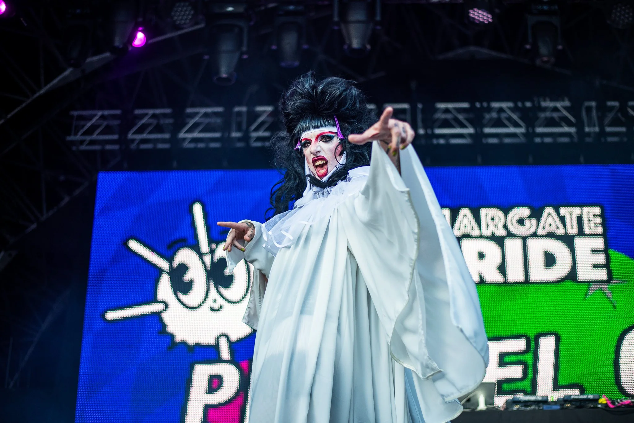 Performer in dramatic costume on stage at Margate Pride event with colorful digital backdrop, spotlights overhead.