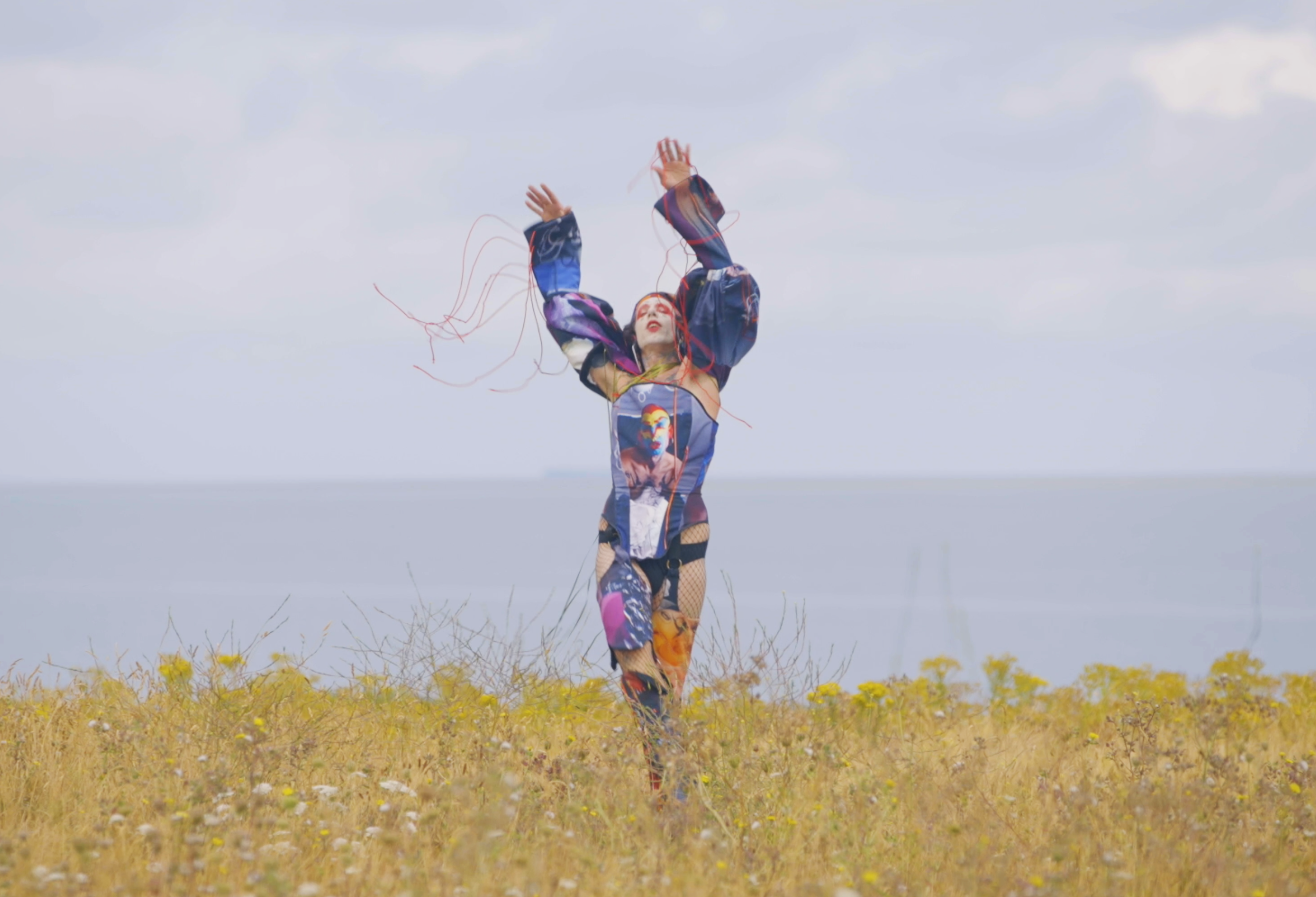Person in colorful, artistic costume standing in a field by the ocean, arms raised.