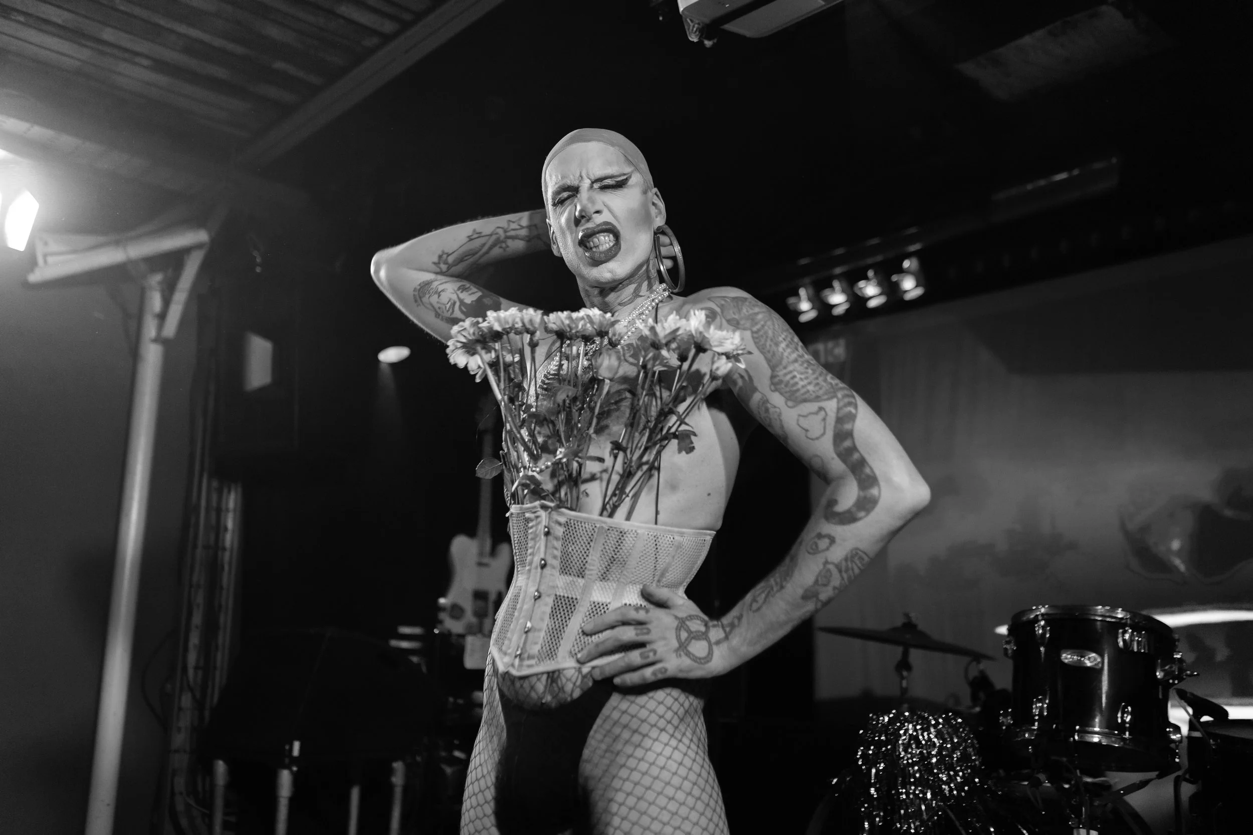 Performer wearing a corset filled with flowers and fishnet stockings on stage with musical instruments in the background.