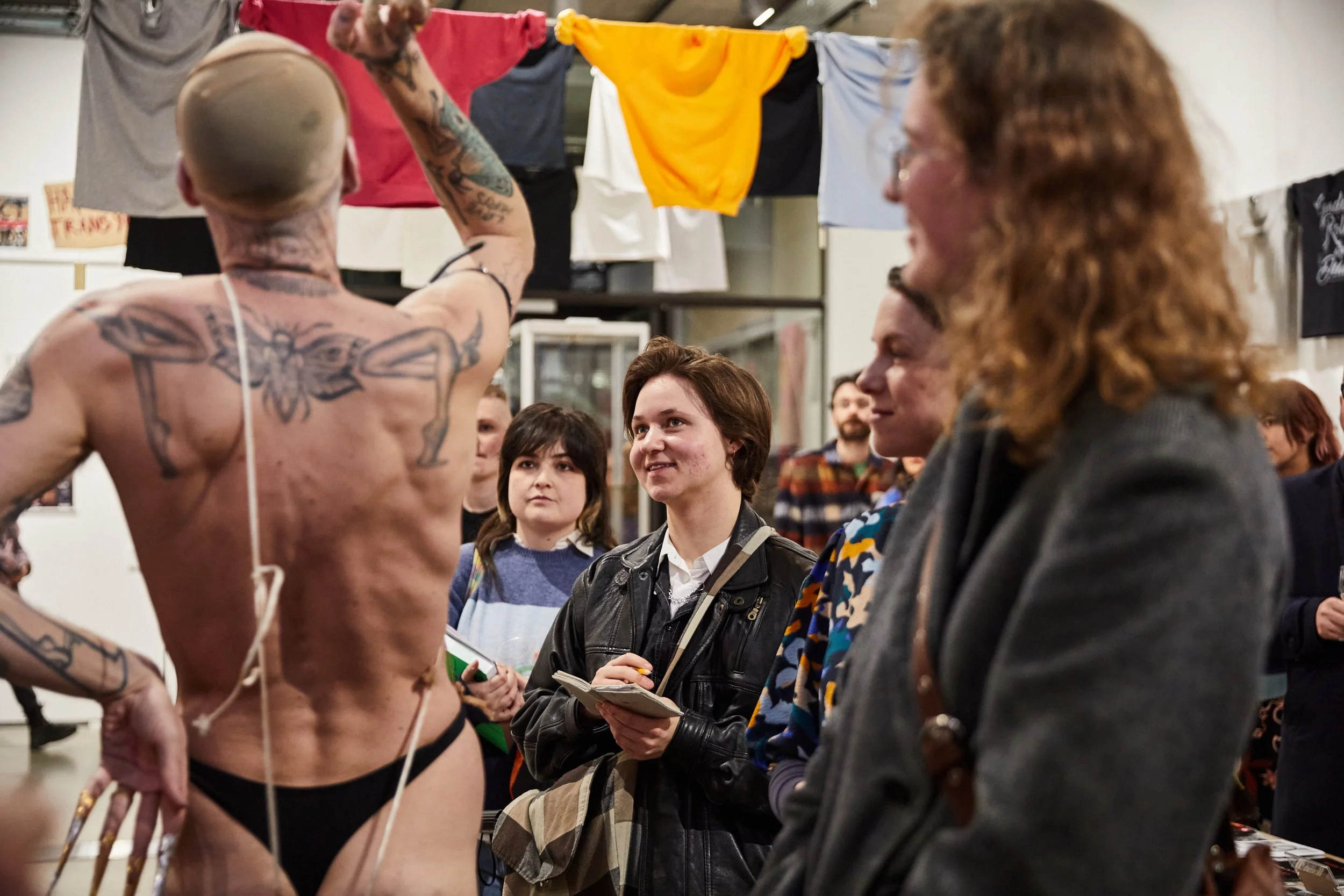 A shirtless man with tattoos and a shaved head demonstrating at an event, surrounded by a diverse group of people watching and taking notes.