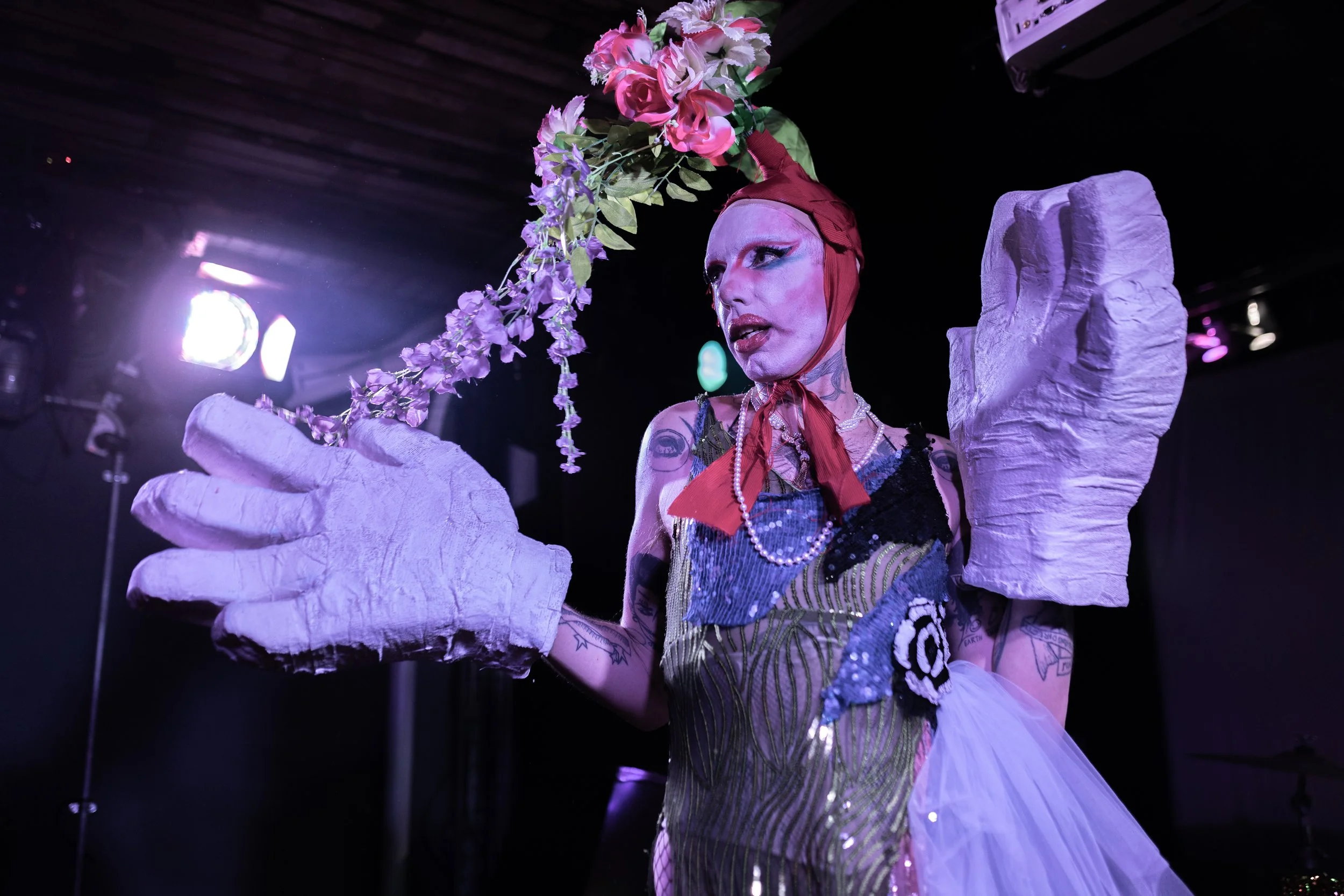 Person in colorful costume with large hand props and floral headpiece, performing under stage lights.