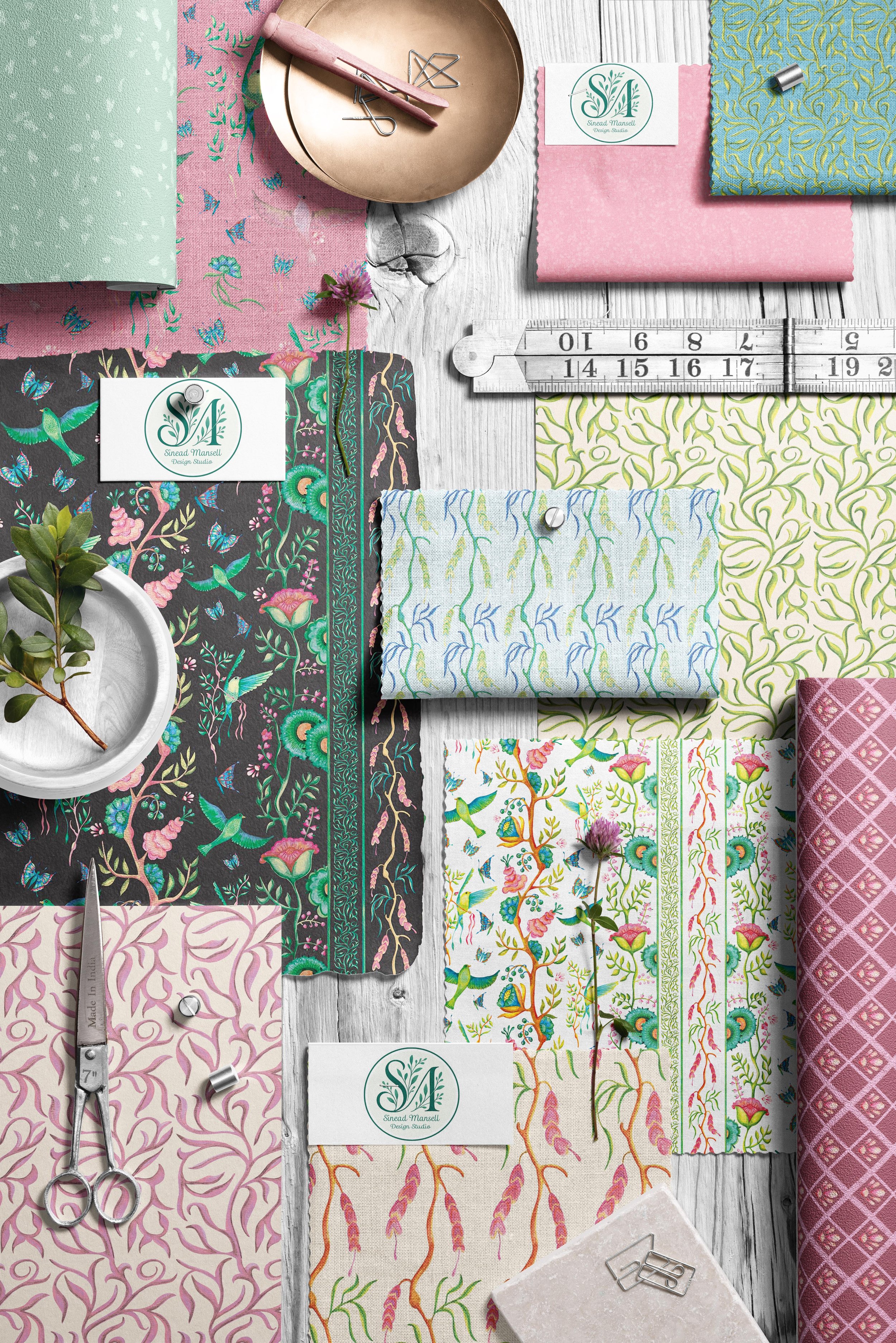 Various patterned fabrics with floral and nature-inspired designs are arranged on a white wooden surface. Items such as scissors, a measuring tape, fabric clips, a small dish with greenery, and a business card with a logo are included in the arrangem