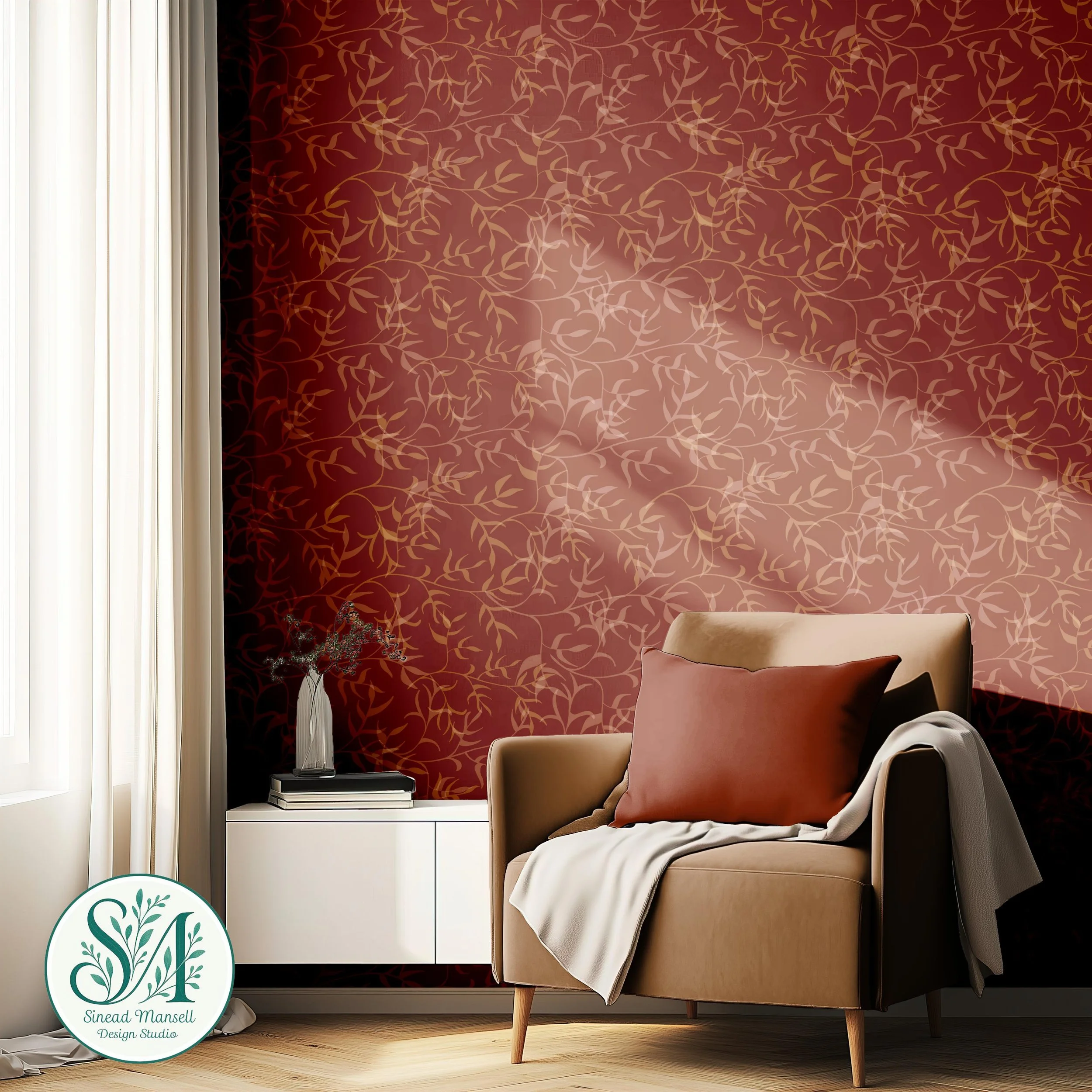 A cozy living room corner with a beige armchair, soft pink and beige pillows, a throw blanket, a white side table with books and a small plant, and a dark red wall with a gold vine pattern. Light filters through white curtains on the left.