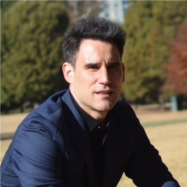 A man with dark hair and a slight smile, wearing a dark shirt, outdoors in a park with trees in the background.