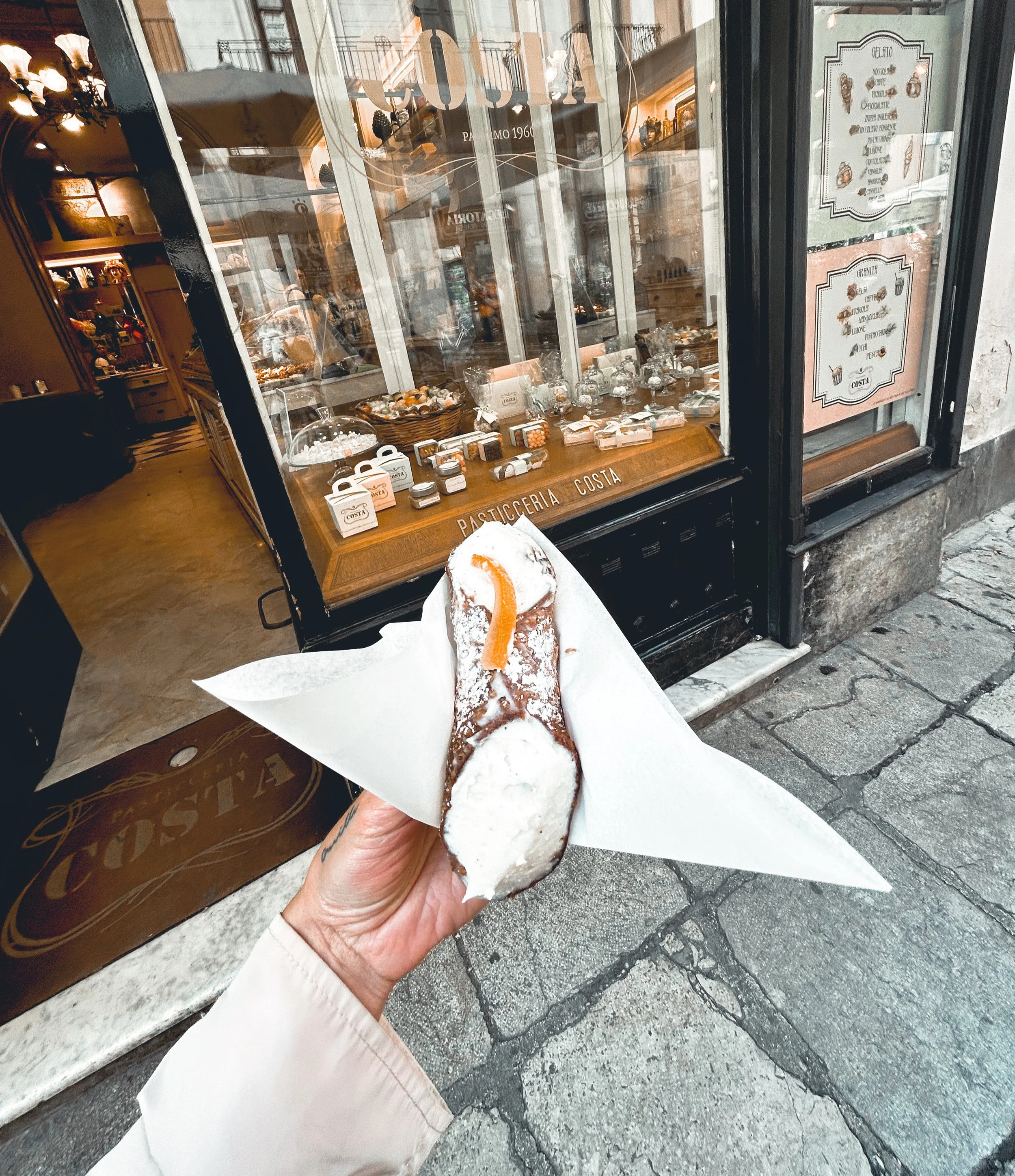 Hand holding a cannoli with powdered sugar and an orange peel in front of a pastry shop window decorated with sweets and signs.