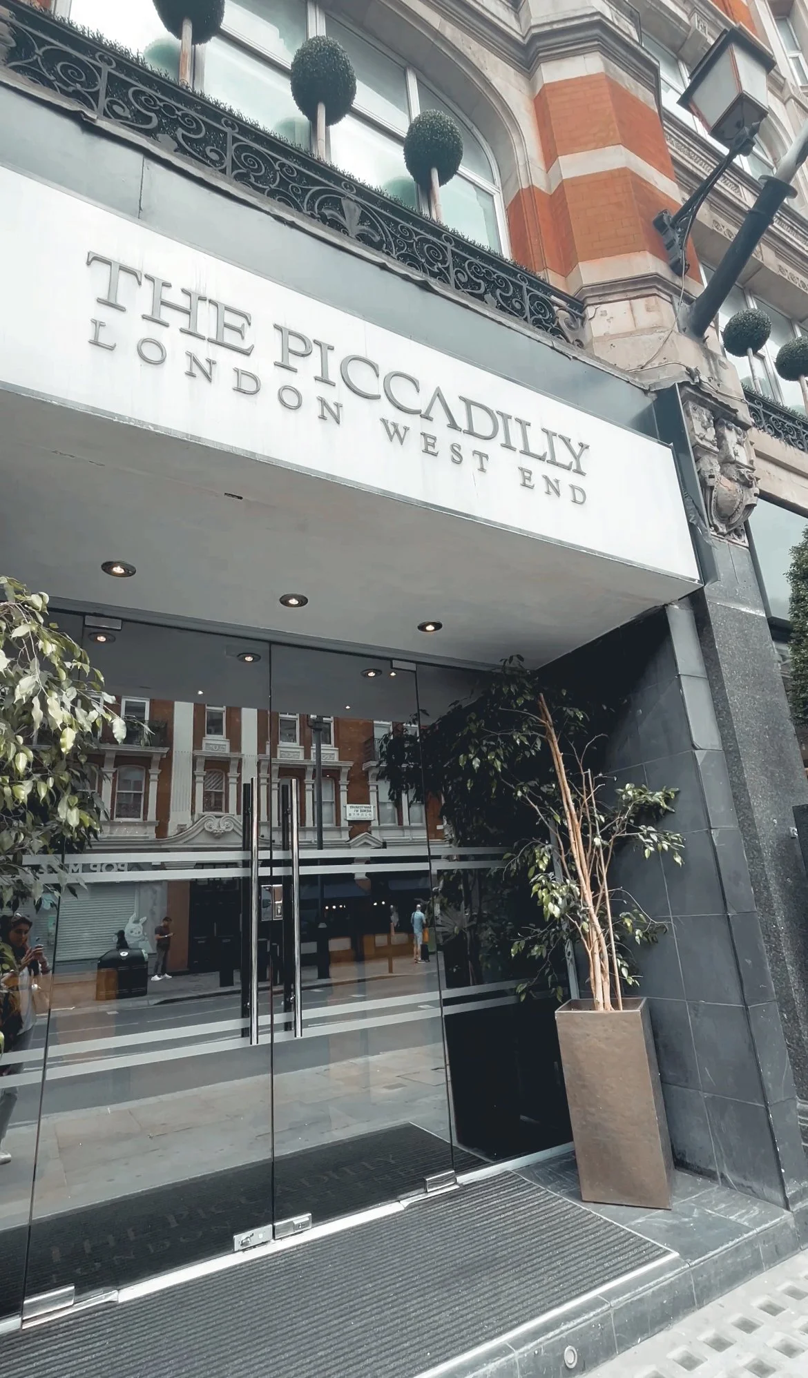 Exterior view of The Piccadilly London West End hotel showing glass doors, plants, and the hotel sign on the building.